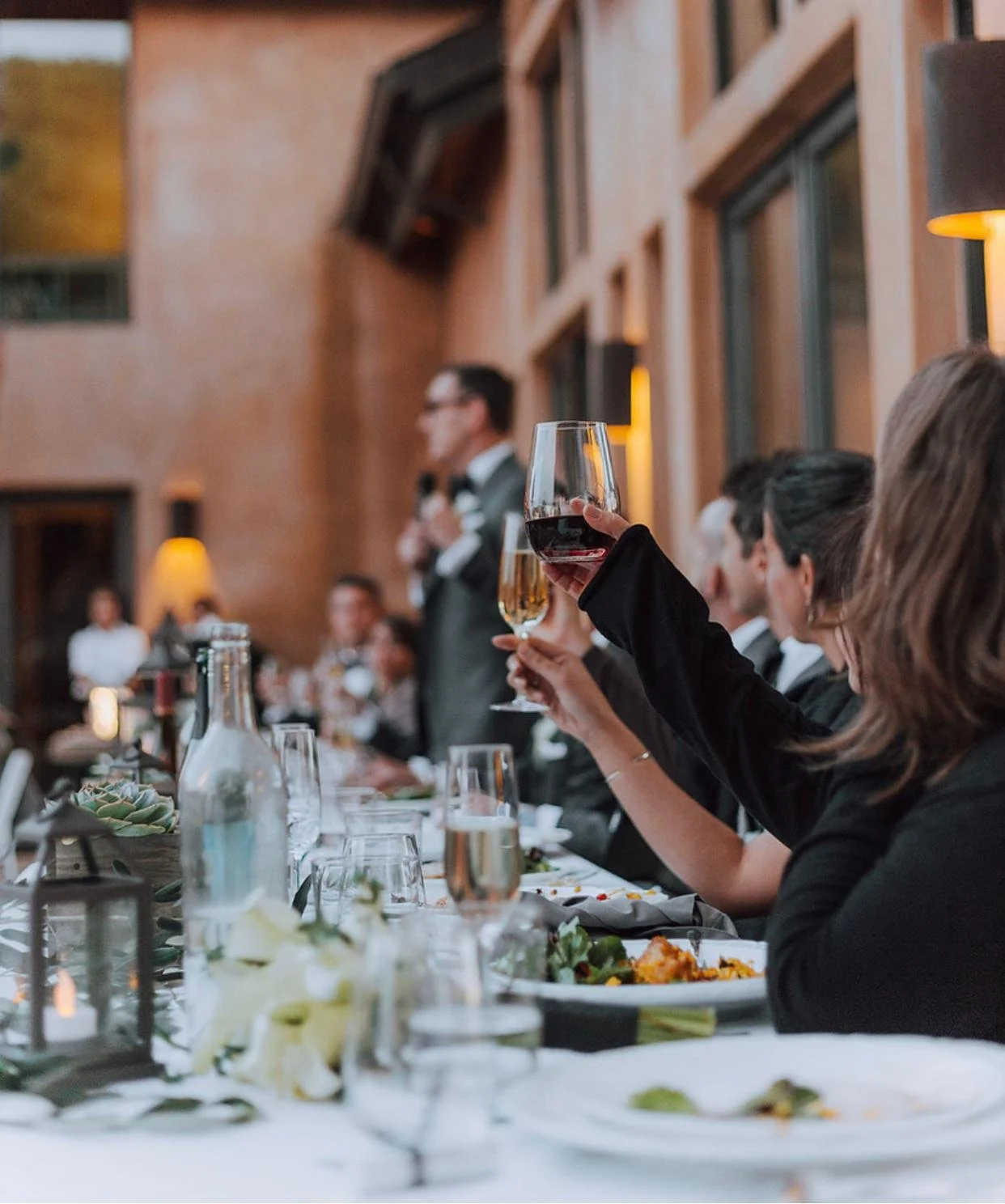A group of formally dressed people at a dinner event, raising glasses of wine and champagne for a toast, with a speaker standing in the background.