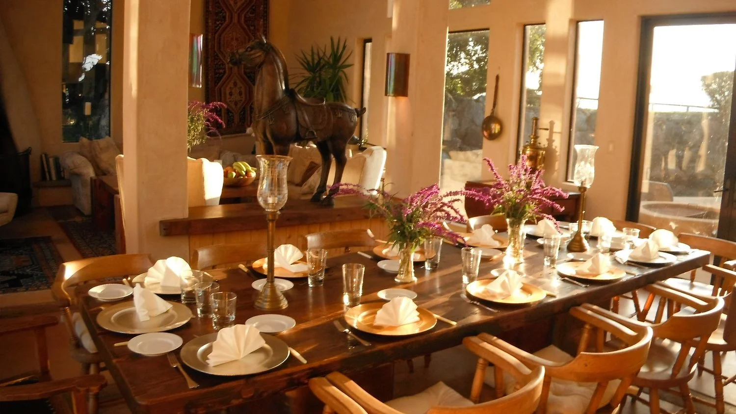 Dining room with a long wooden table set with gold and white plates, white napkins folded in a fan shape, glassware, and candles. Pink flowers in vases are on the table. In the background, a wooden horse sculpture, a plant, a tapestry, and windows le