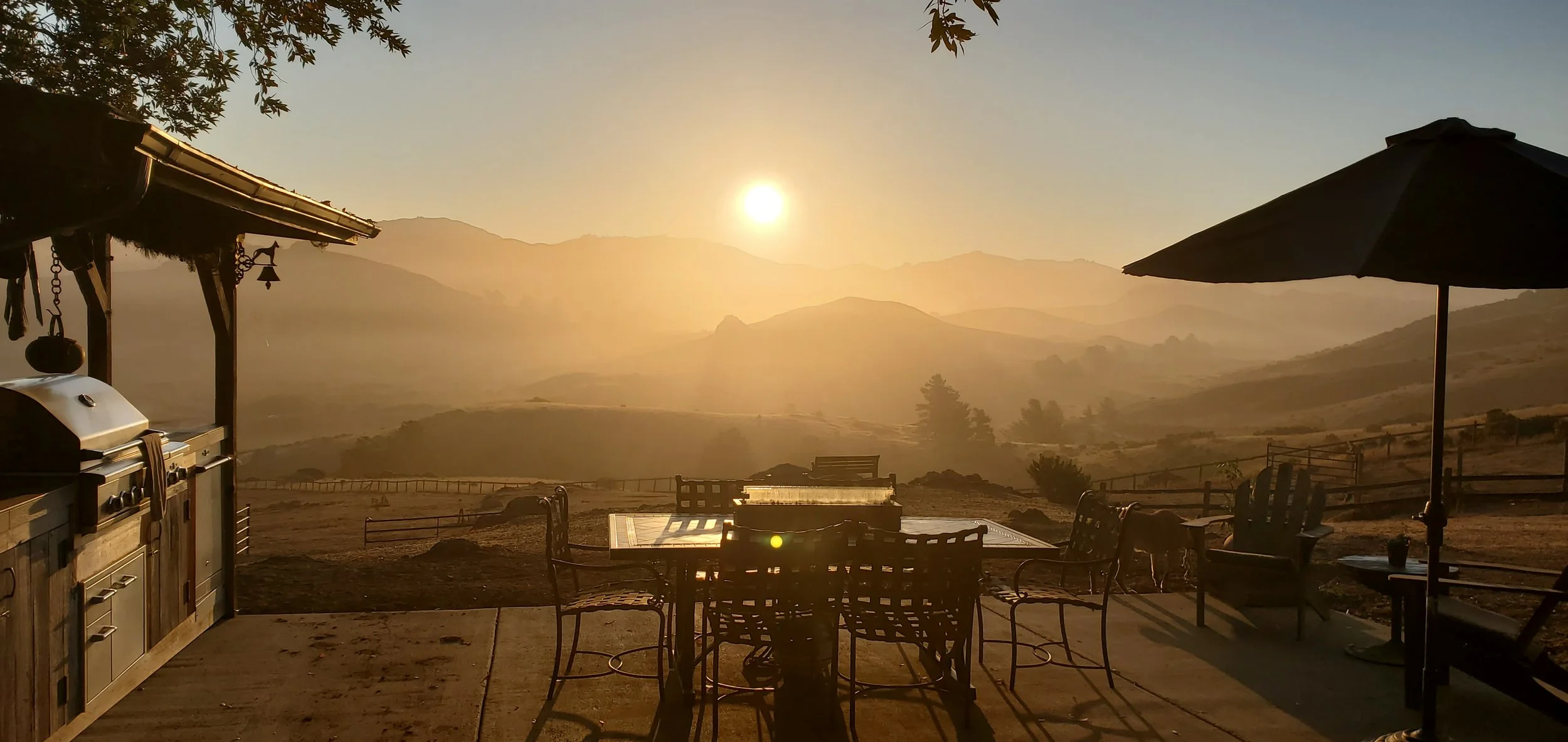 Sunset over a mountainous landscape viewed from a patio with outdoor furniture, a grill, and a large umbrella.