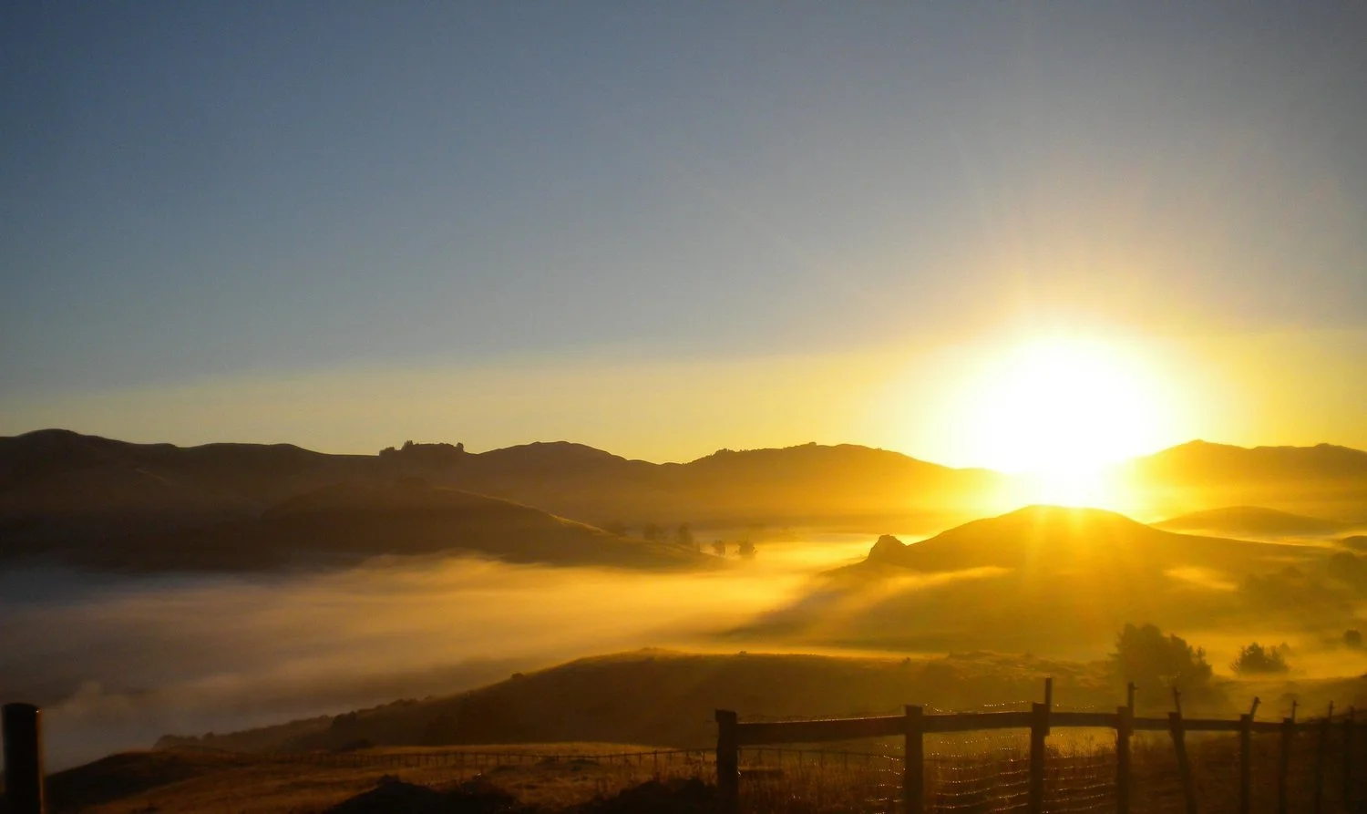 Sunrise over rolling hills with fog and a wooden fence in the foreground.