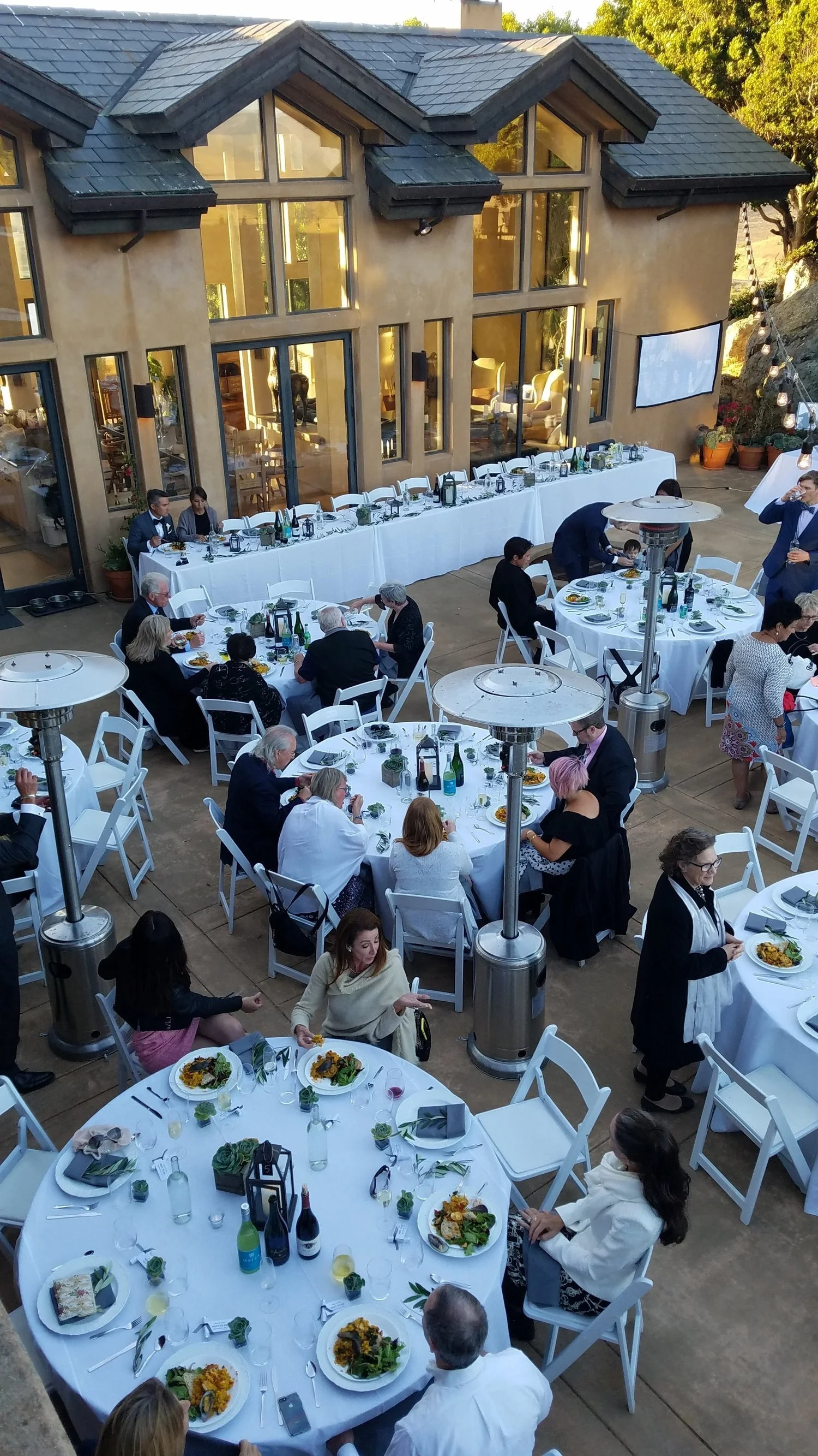 An outdoor dining event with multiple round tables covered with white tablecloths, set with plates of food, glasses, and napkins. People are seated and enjoying their meals, with some standing and conversing. There is a long rectangular table with a 
