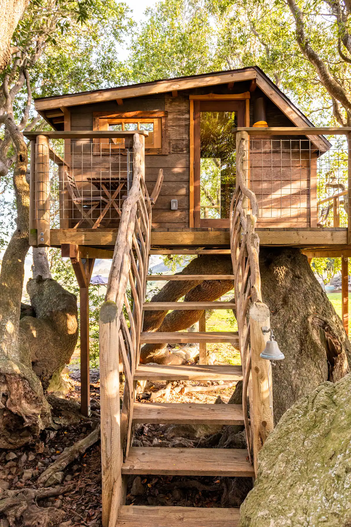 A wooden treehouse built among the branches of a large tree, accessed by a set of wooden stairs with decorative twisted handrails. The house has a small window, a door, and a balcony with a railing, all surrounded by lush green leaves.