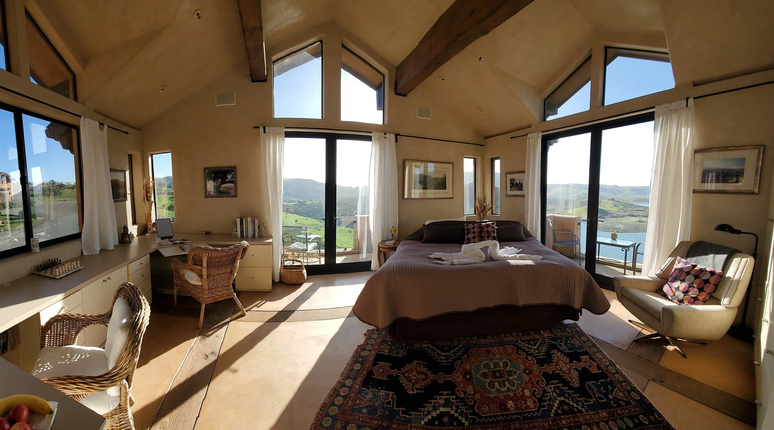 A cozy bedroom with large windows and glass doors showing a scenic green landscape and hills outside. The room has a bed with a folded towel swan on it, a sitting area with a cream armchair and a decorative pillow, a desk with a computer, and various framed artwork on beige walls. Sunlight streams into the room, illuminating a patterned rug on the floor.