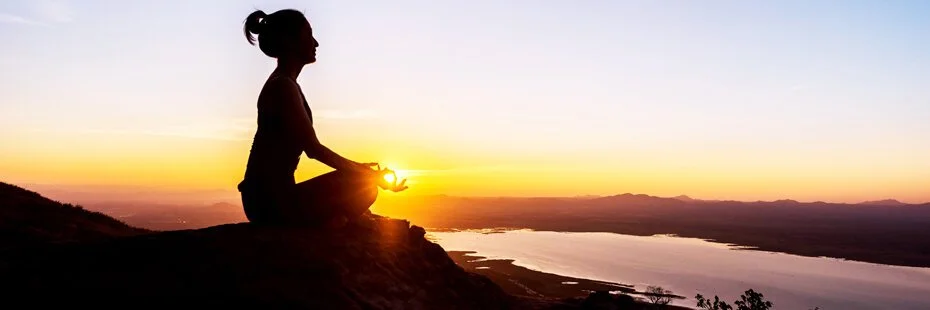 Silhouette of a woman meditating on a hilltop during sunset near a body of water.