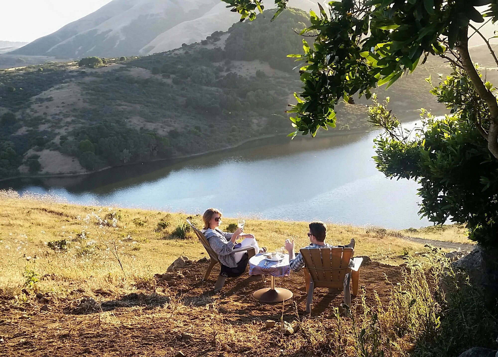 People relaxing and enjoying drinks on a hillside with a water body and mountains in the background, during a sunny late afternoon.