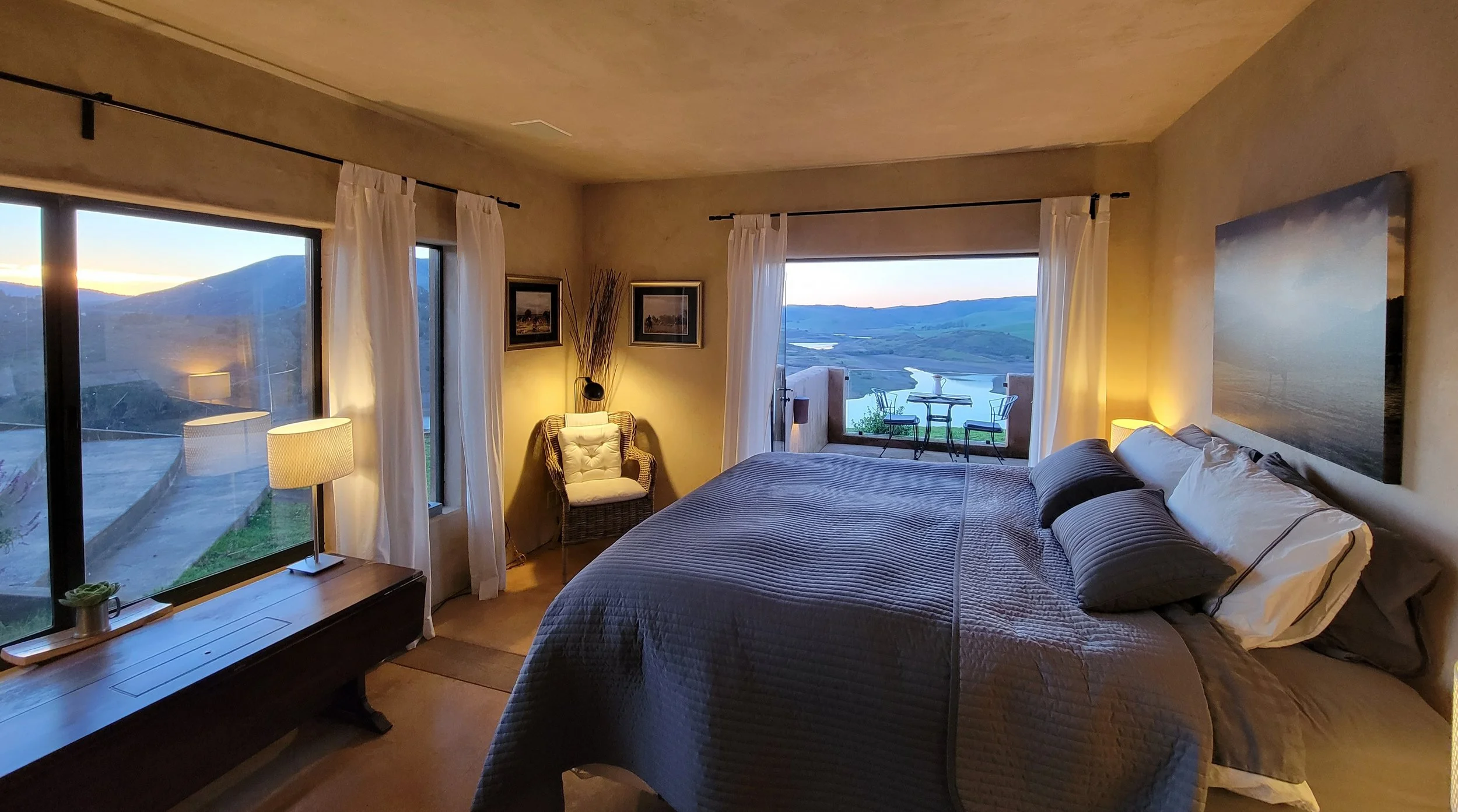 A cozy bedroom with large windows showing mountains, a lake, and a balcony with outdoor furniture. The room has a bed with pillows, lamps, framed pictures, and a wicker chair with a tufted cushion.