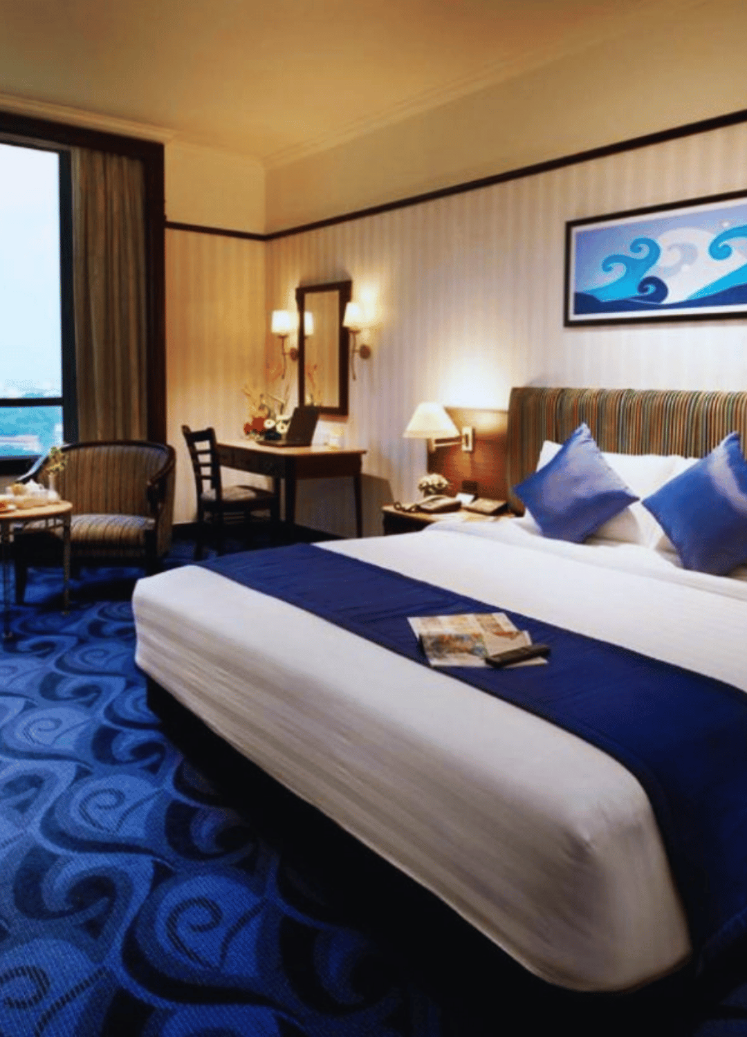 A hotel room with a large bed, blue pillows, a blue bed runner, and a city view from the window. There's a sitting area with a striped armchair, a small table, and a desk with a mirror, lamps, and decorative items.