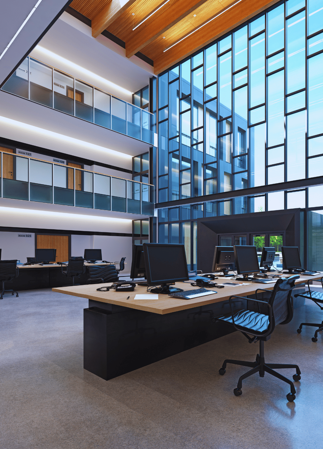 Modern open-plan office with multiple desks, computer workstations, and black office chairs, featuring a large glass wall and upper balcony in a high-rise building.
