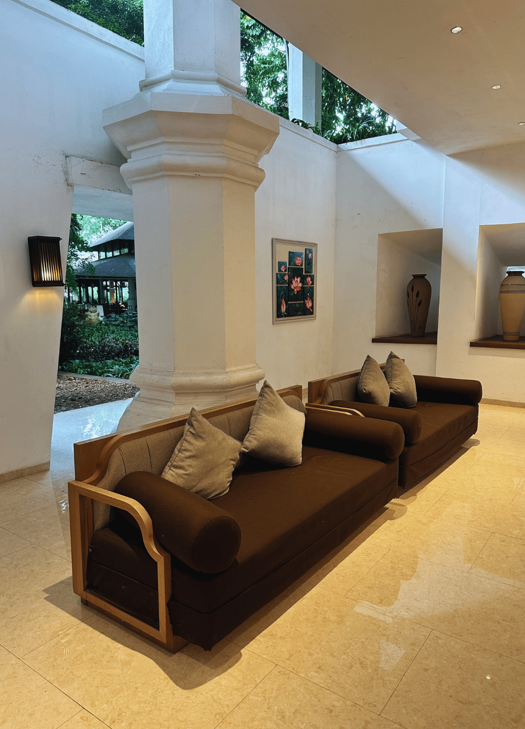 Interior lobby area with two brown sofas, beige throw pillows, white walls, a large white pillar, and a picture frame on the wall. Natural light and greenery visible outside through open sections.