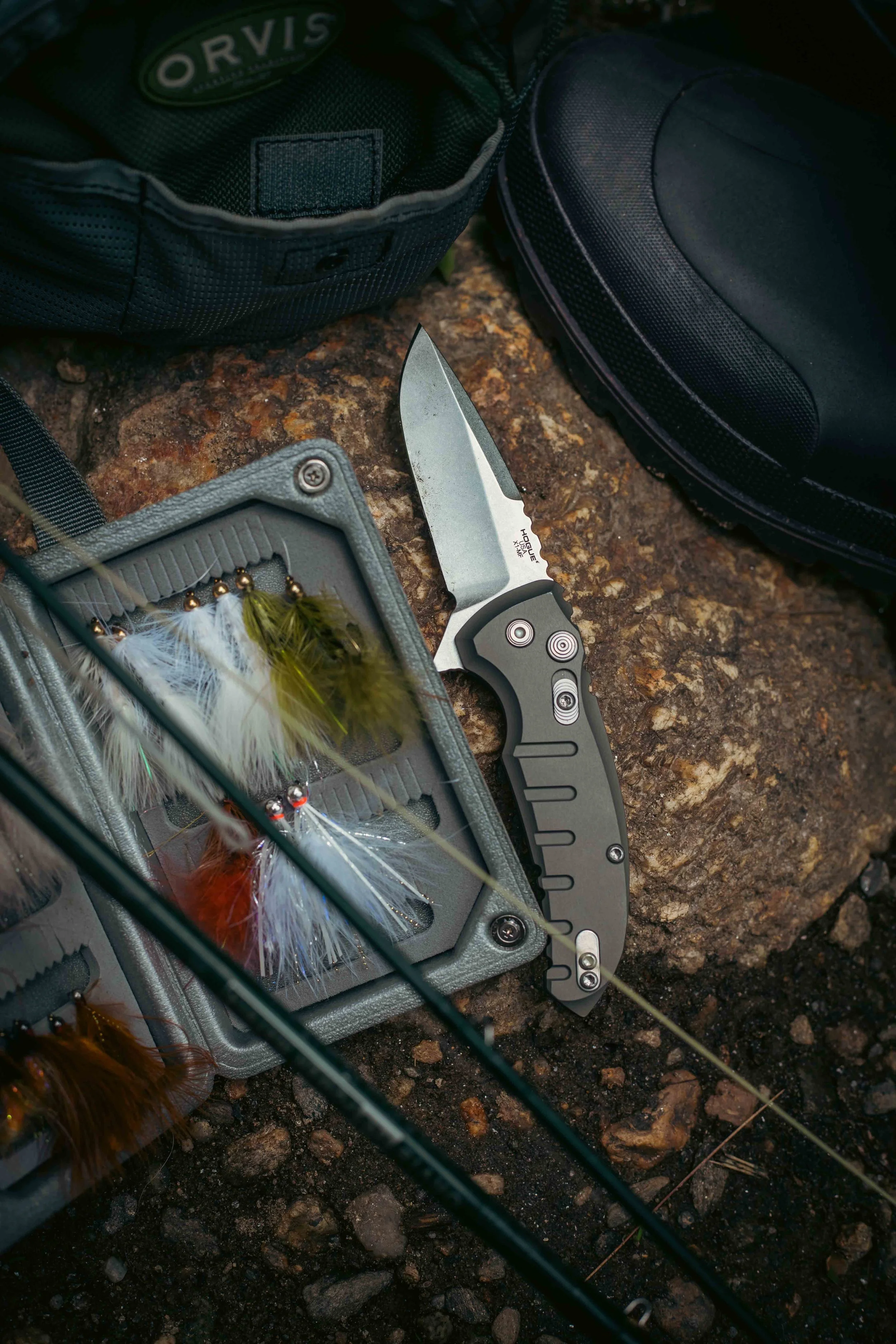 Fishing gear including a folding knife, fishing lures with feathers, and a tackle box on rocky ground, with a black backpack and black bag nearby.