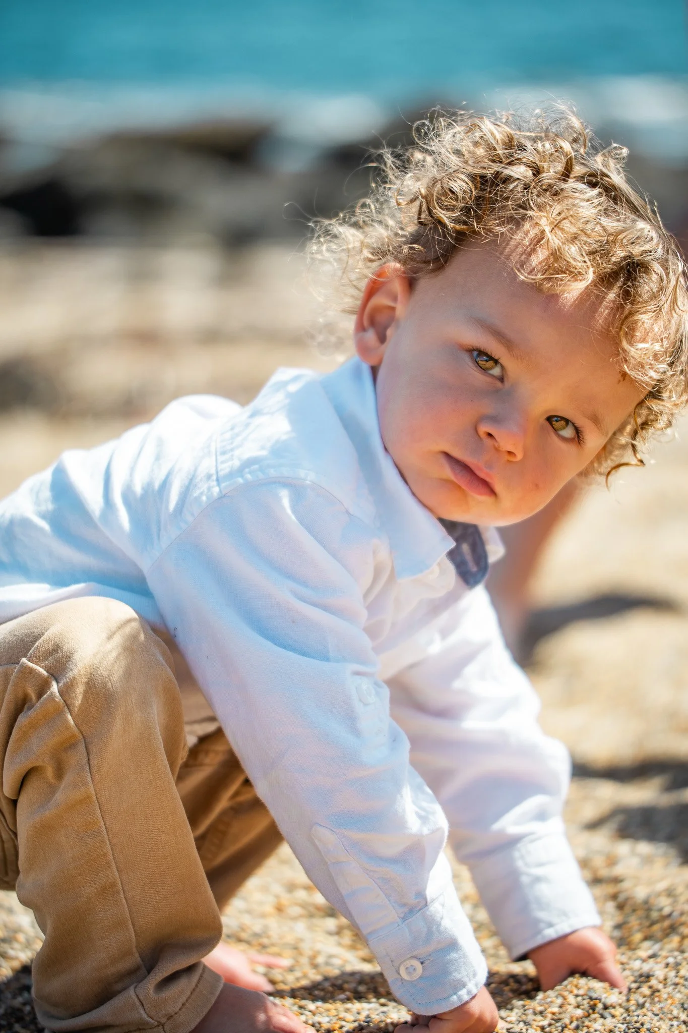 A young boy with curly blond hair and light skin playing on a sandy beach, crawling on his hands and knees with a focused expression.