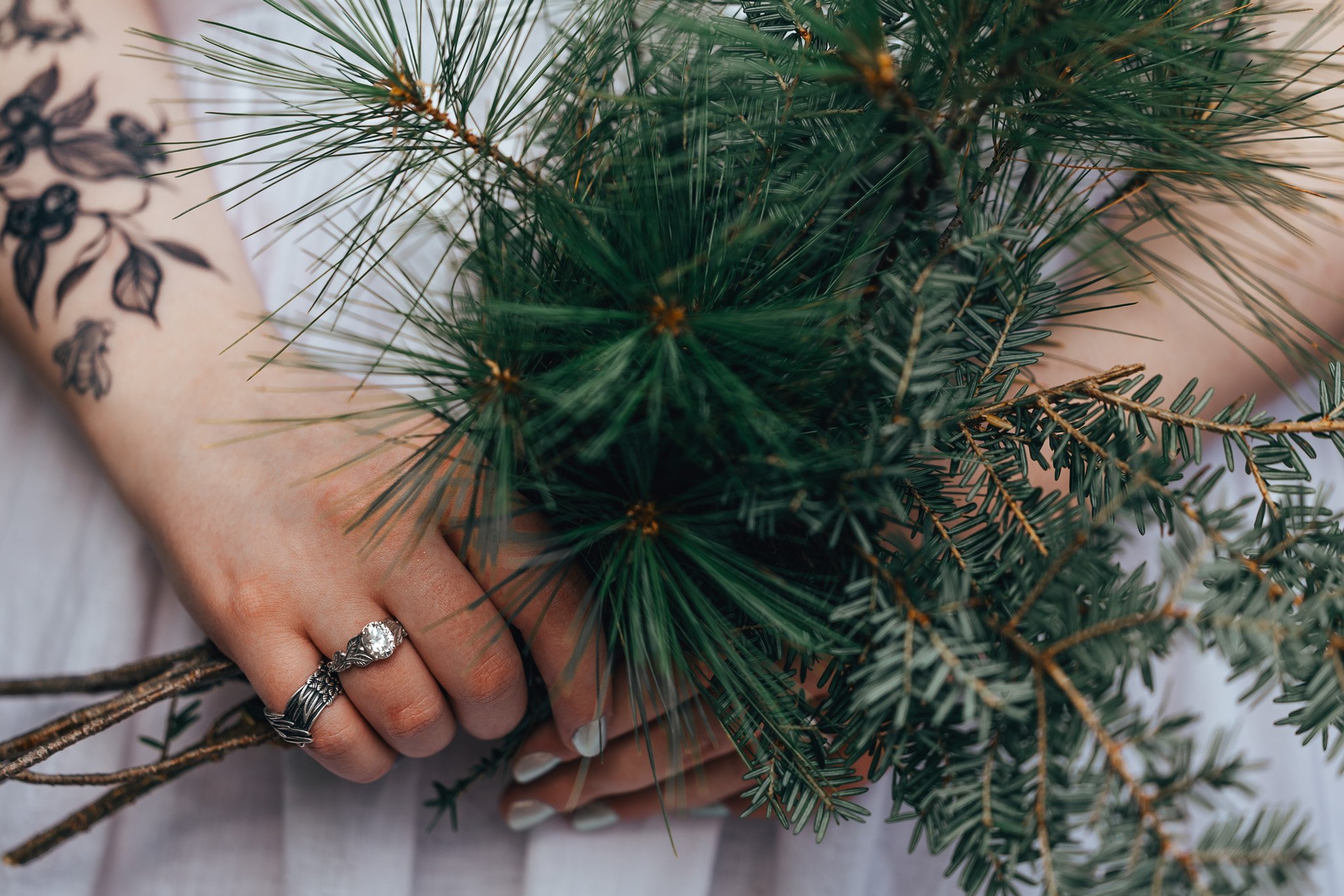 A person's hand holding pine branches, with a tattoo of leaves on the arm and a diamond ring on the finger.