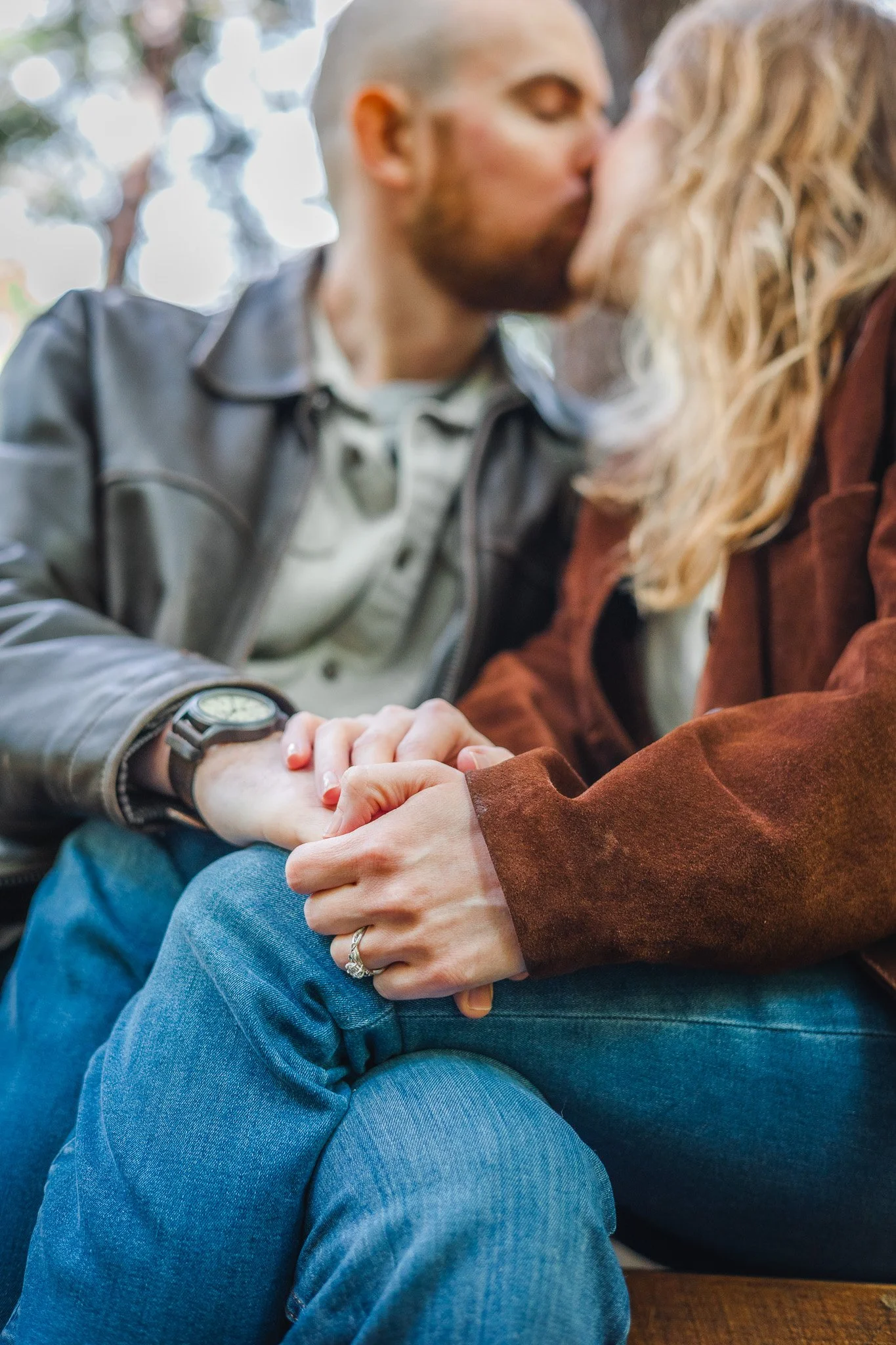 A man and woman holding hands and sharing a kiss, close-up of their hands and faces, with blurred background of trees.