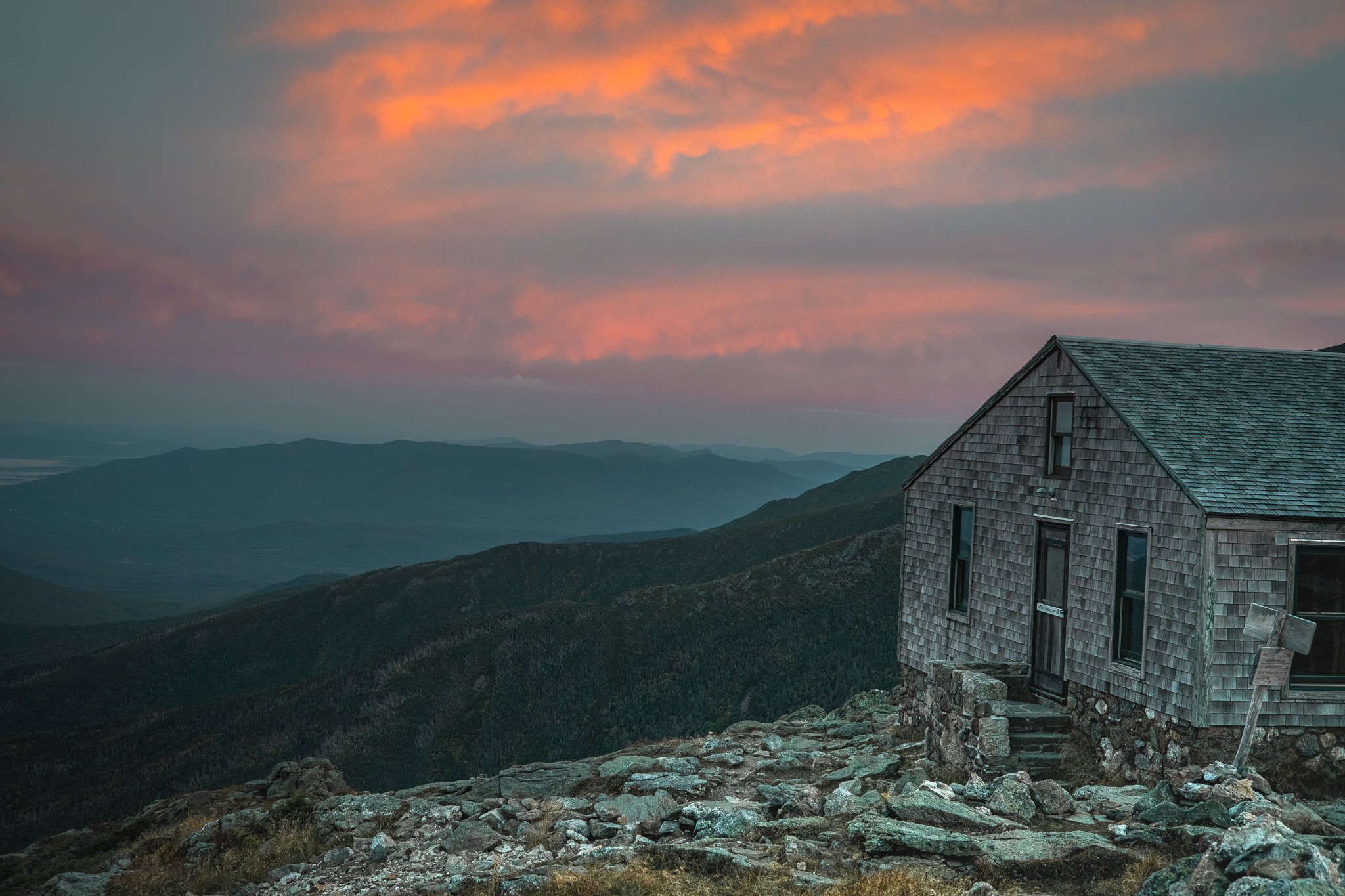A rustic wooden house with a shingled roof is situated on a rocky hillside under a colorful sky at sunset, with rolling mountains in the background.
