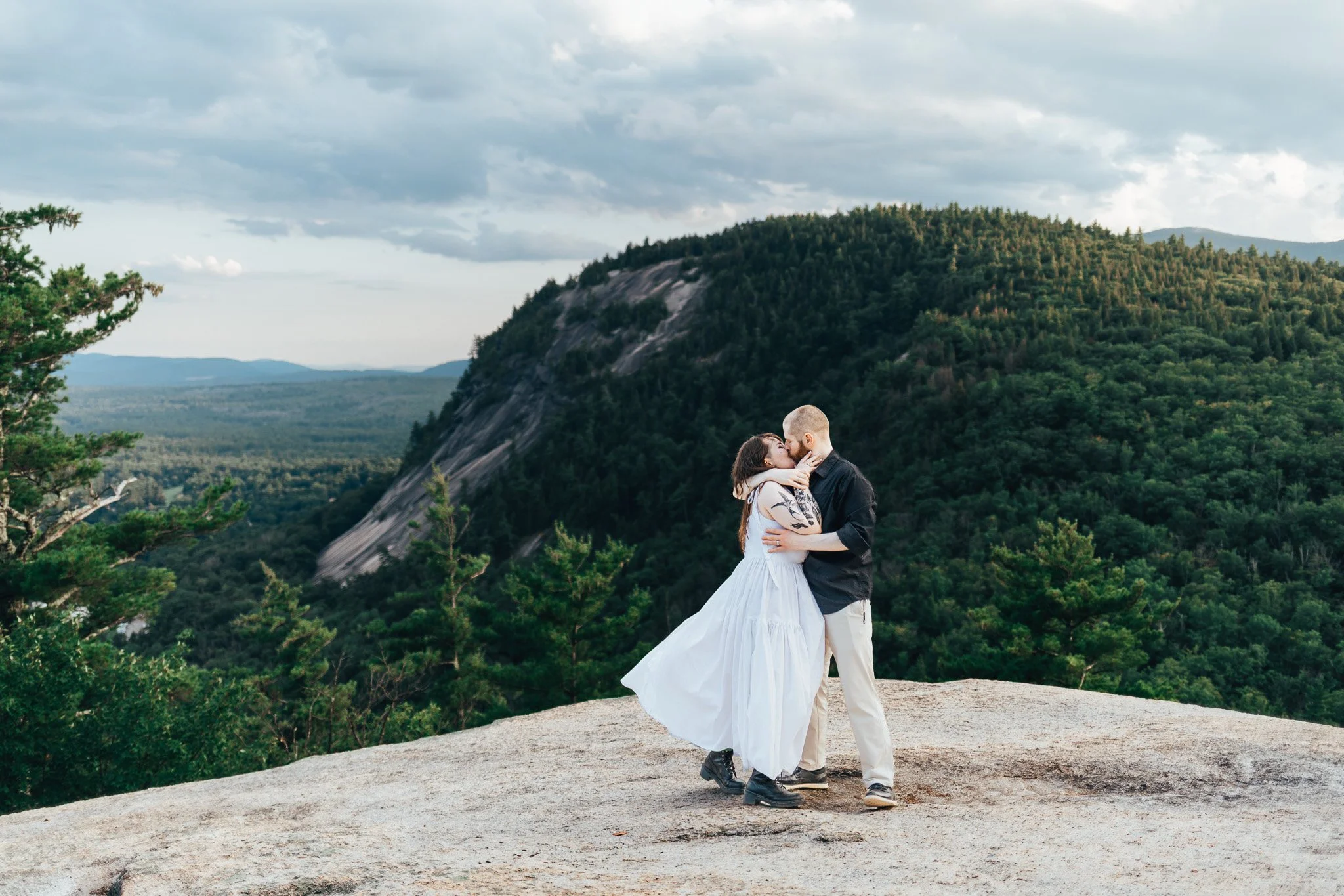 A couple sharing a kiss outdoors on a rocky surface with lush green mountains and a cloudy sky in the background.