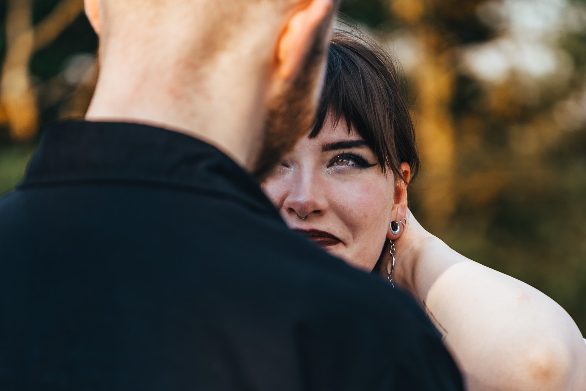 A woman with dark hair and large earrings looking at a man with red hair, who is turned away from the camera, outdoors with a blurred, autumn-colored background.