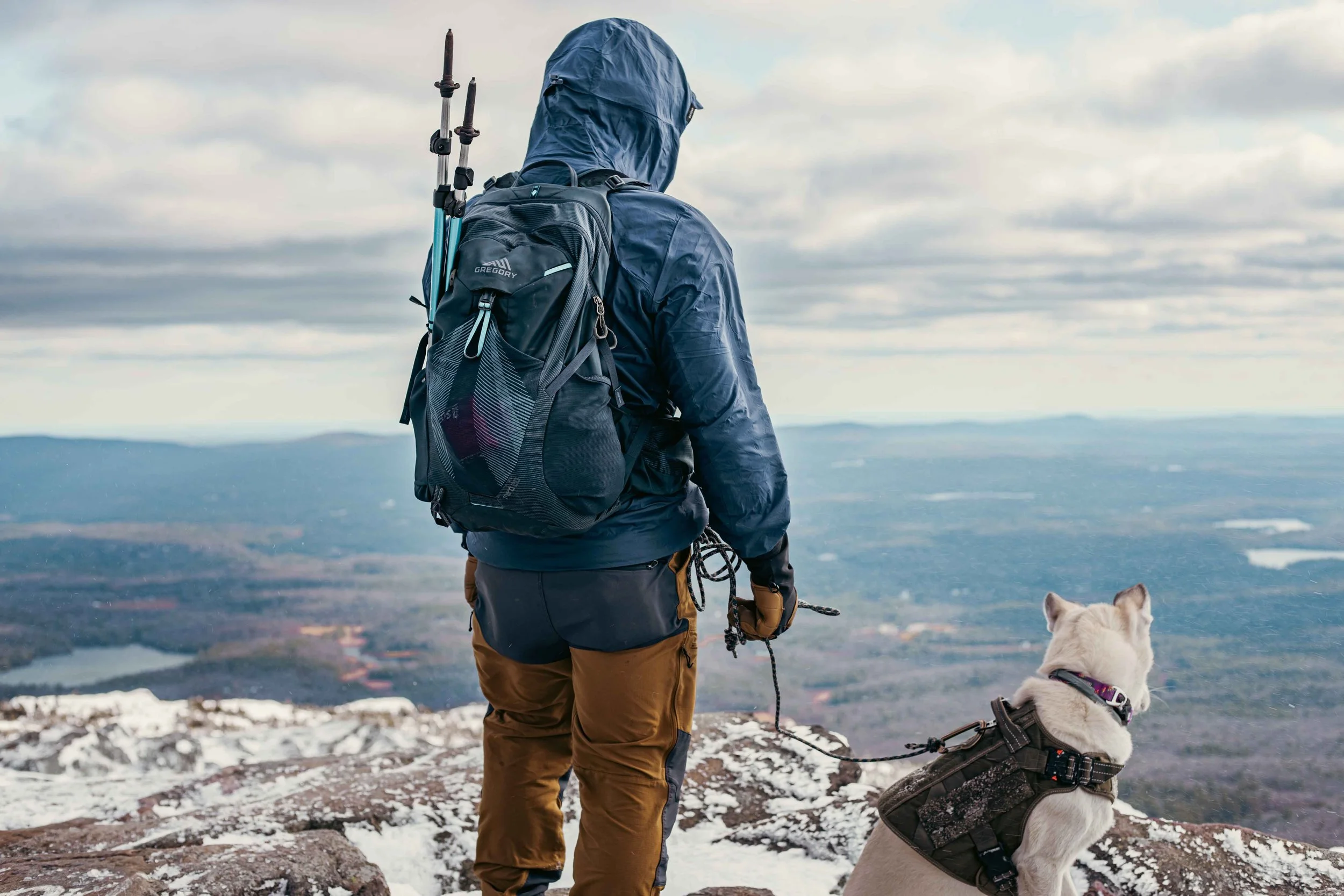 A person wearing a blue jacket and brown pants hiking with a backpack and trekking poles, holding a leash attached to a white dog in a snow-covered mountainous landscape during daytime.