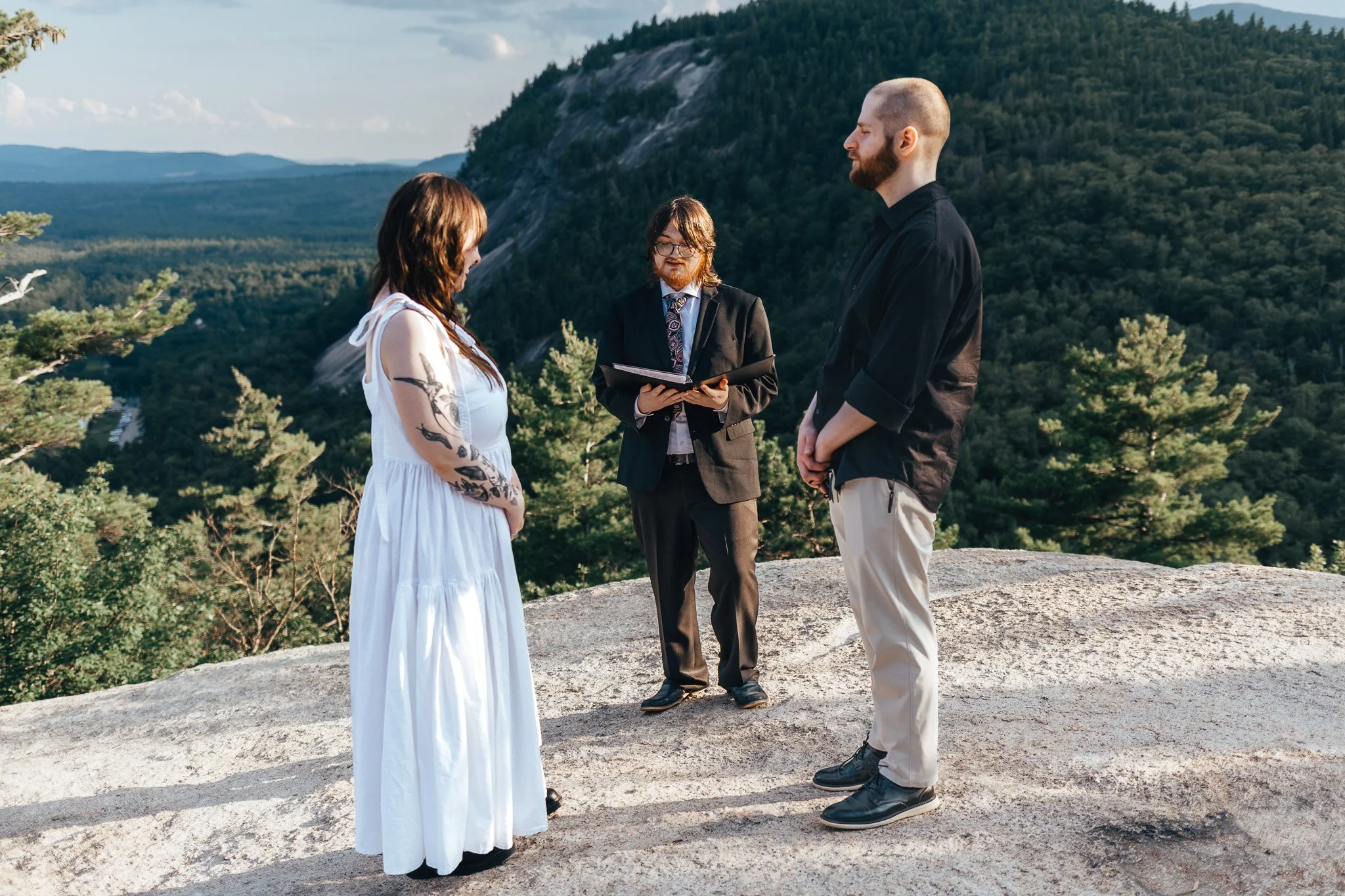 A wedding ceremony taking place outdoors on a rocky surface with a forested mountain background. A woman in a white dress with tattoos on her arms is standing across from a man in a black shirt and white pants. An officiant in a black suit and patterned tie stands between them, holding a book.