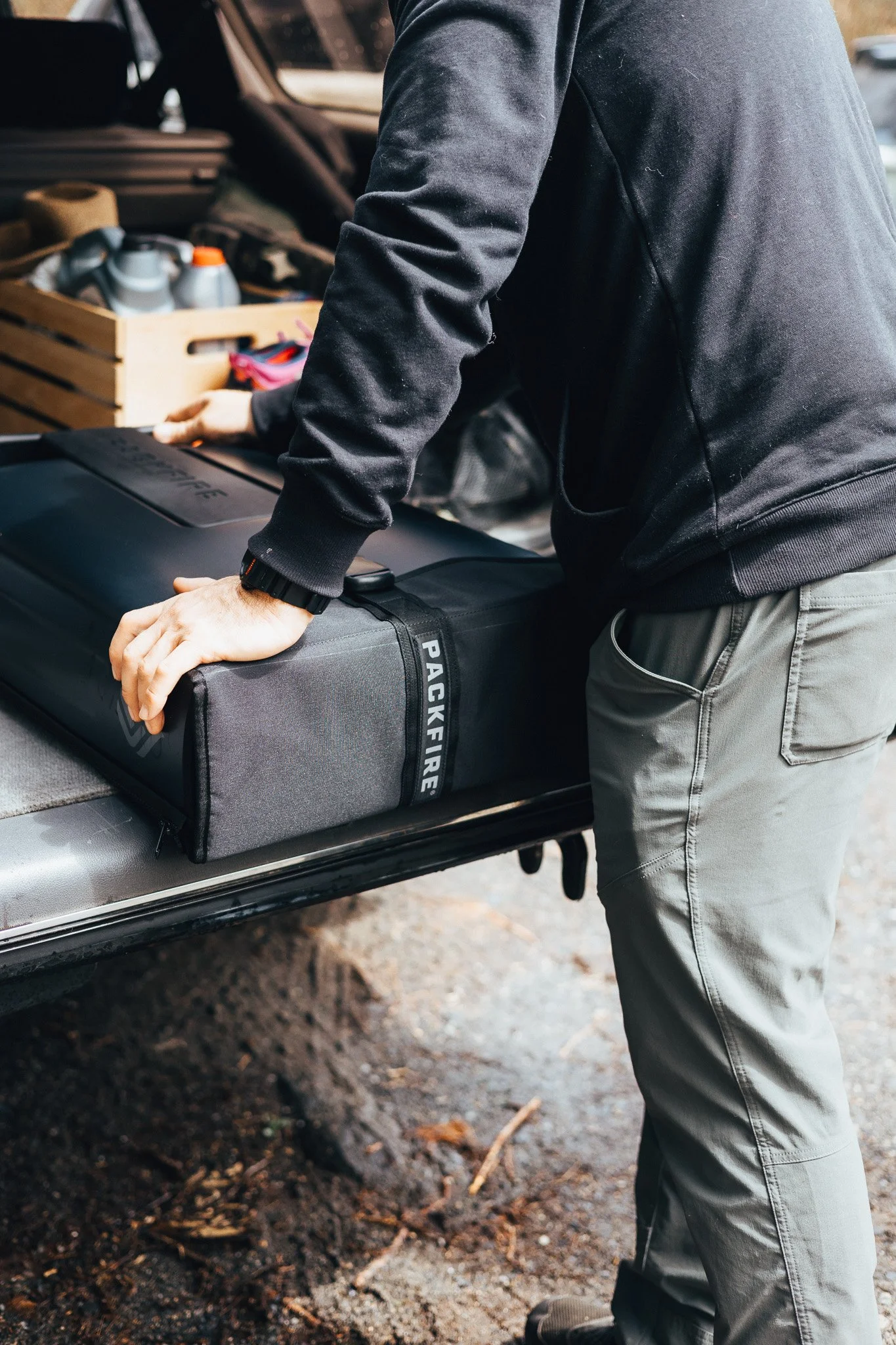 Person in black hoodie loading a toolbox labeled 'PACKFIRE' into a vehicle with a wooden crate containing bottles in the trunk.