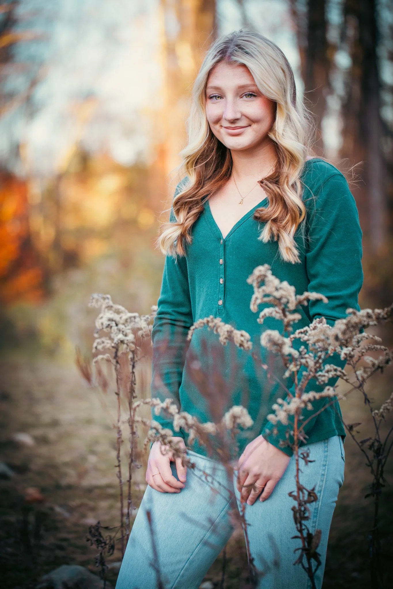 A young woman with blonde wavy hair and blue eyes, standing outdoors in a natural setting with trees and dried plants, wearing a green button-up top and light blue jeans, smiling softly at the camera.