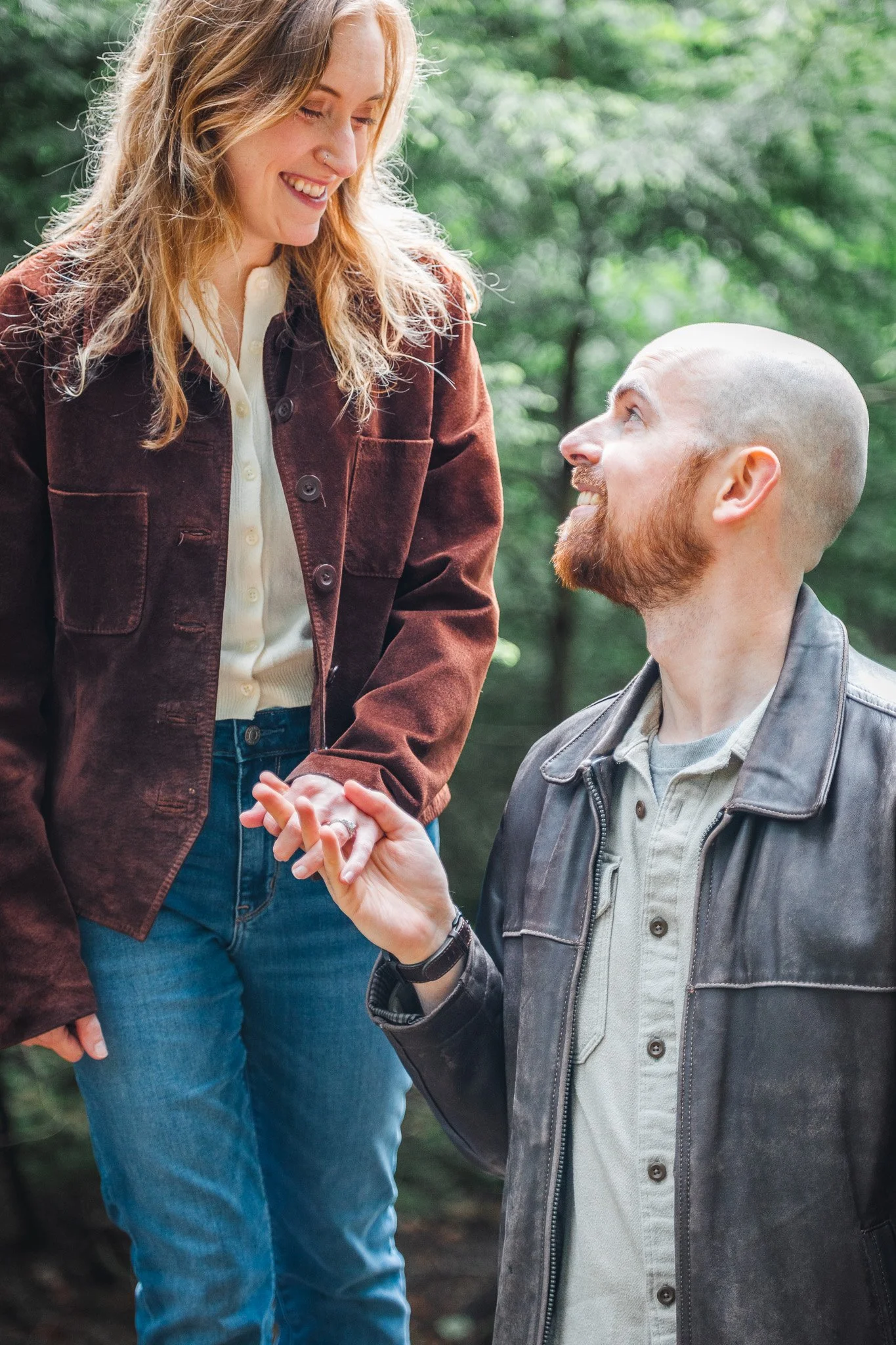A woman with long wavy blonde hair smiling and looking down at a bearded man with a shaved head, who is kneeling and looking up at her. They are holding hands or exchanging a gentle touch. They are outdoors in a forested area with green trees.