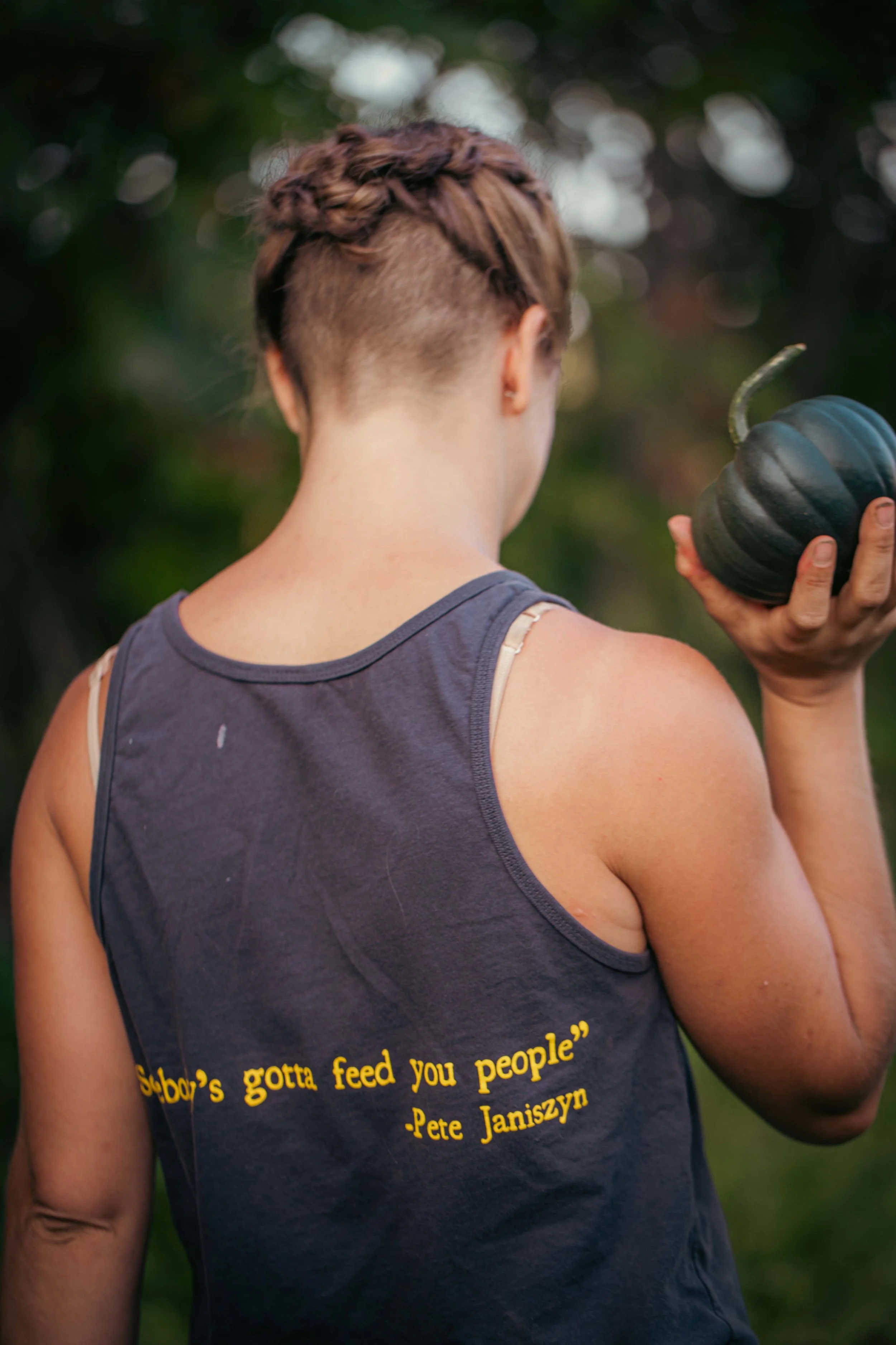 Back of a person with a braided hairstyle, wearing a dark tank top with yellow text, holding a small black pumpkin with a green stem outside with blurred green trees in the background.