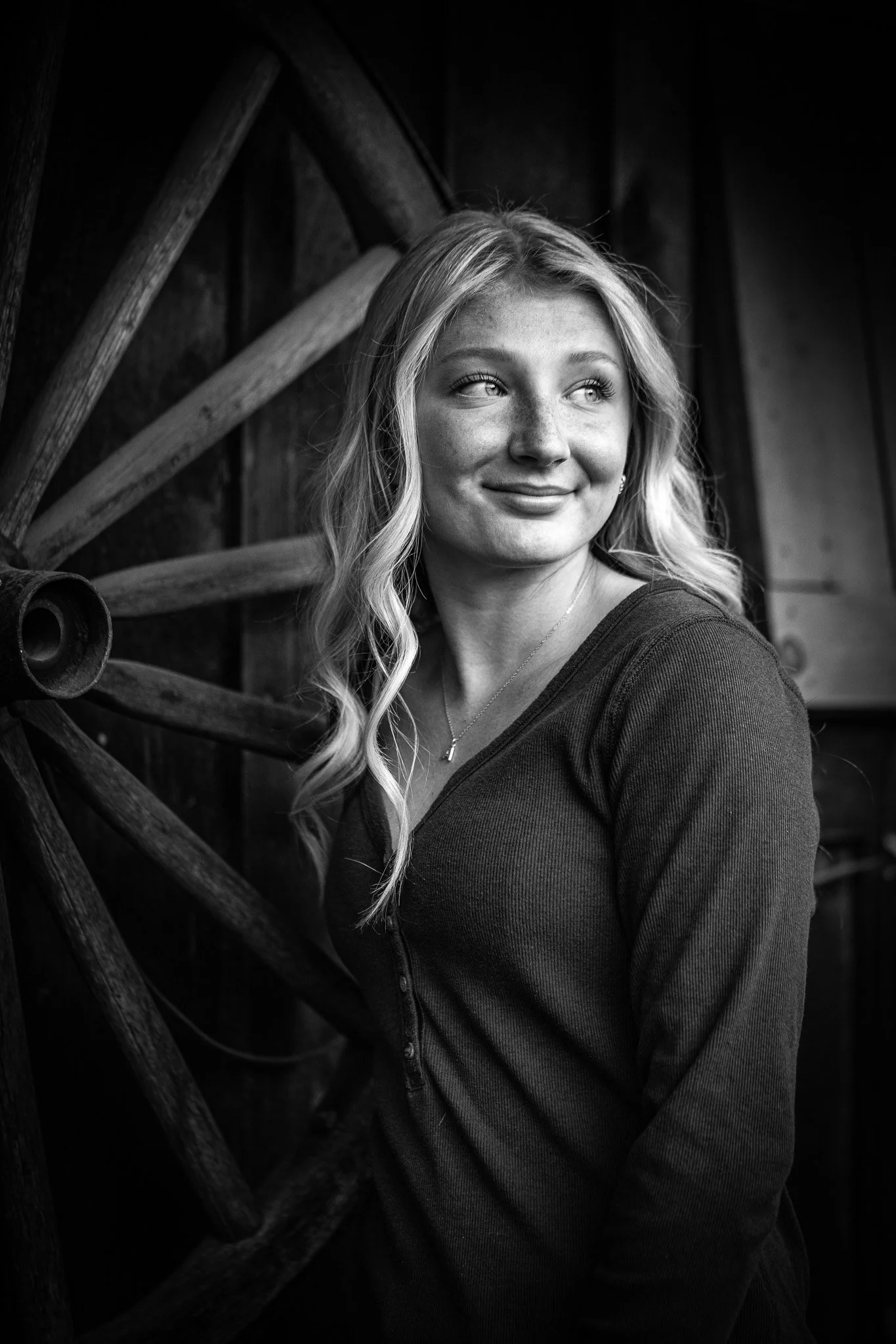 A black and white portrait of a young woman with blonde hair, smiling and looking to her right, standing against a wooden background.