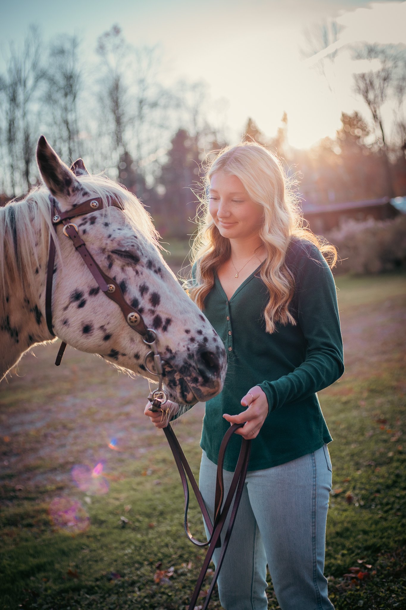 A woman with long blonde hair smiling while holding a leash attached to a white and black speckled horse in a park during sunset, with trees in the background.