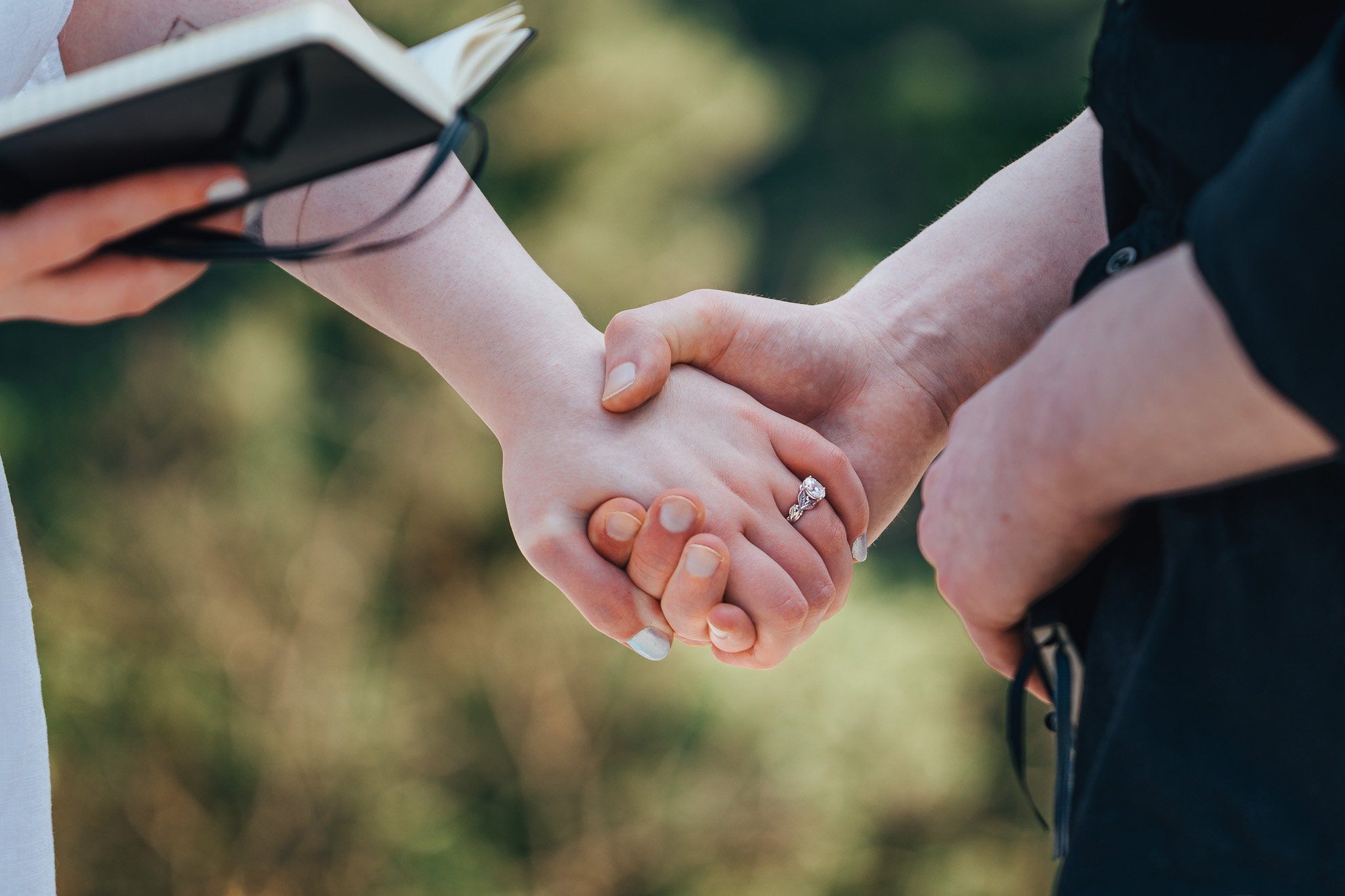 Close-up of two people holding hands during a handshake in an outdoor setting, with the woman wearing an engagement ring.