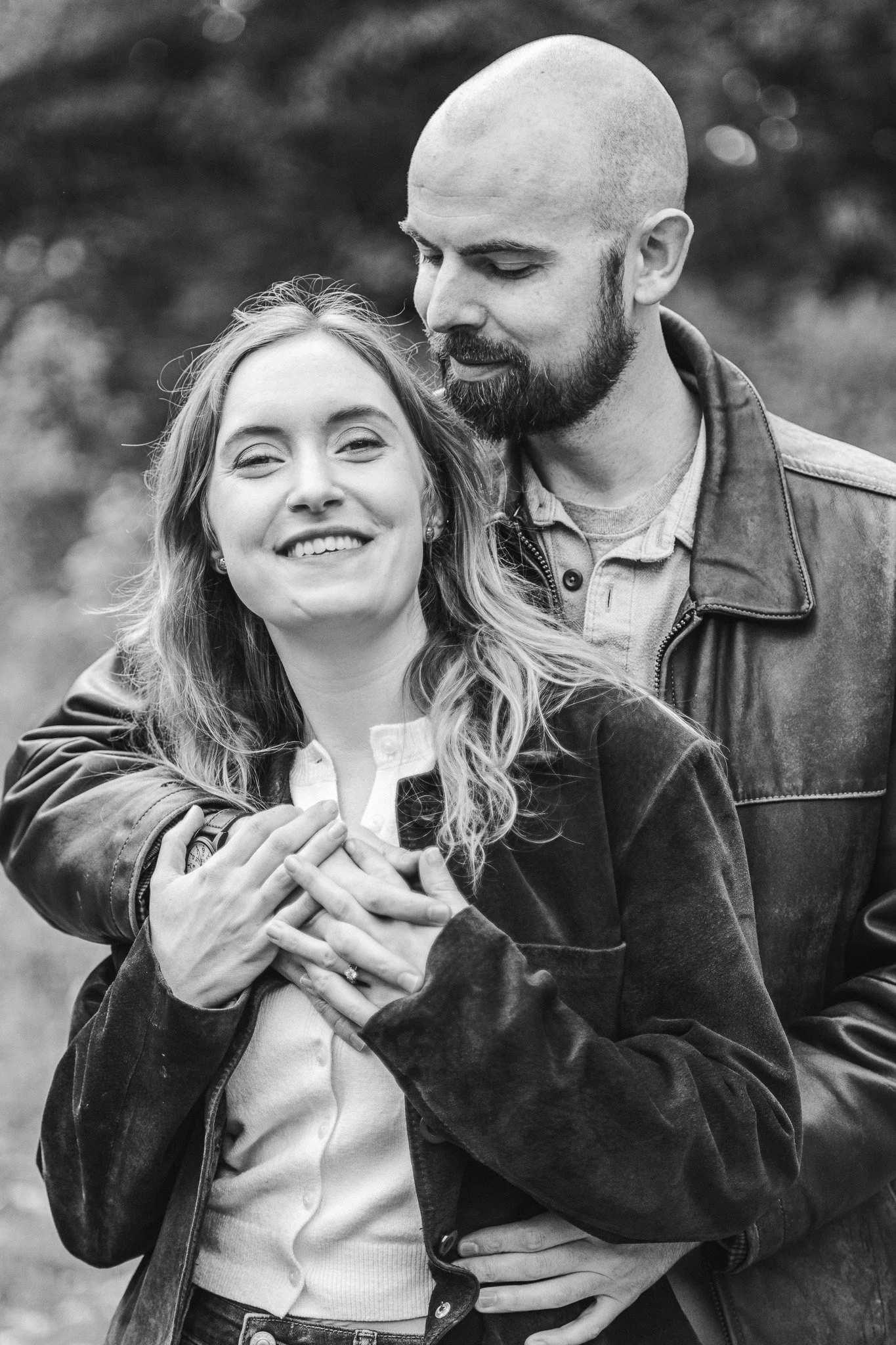 Black and white photo of a smiling woman with long hair embracing a man with a beard and shaved head. They are outdoors, and the woman is wearing a light-colored sweater and a dark jacket. The man is wearing a light t-shirt and a dark leather jacket.