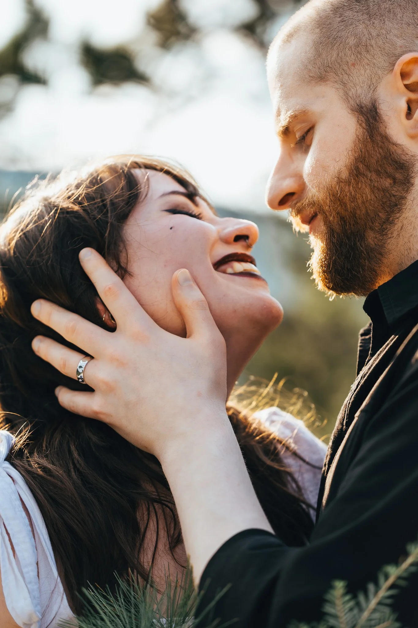 A close-up photo of a couple with eyes closed, face to face, outdoors, with sunlight on their faces, surrounded by greenery.