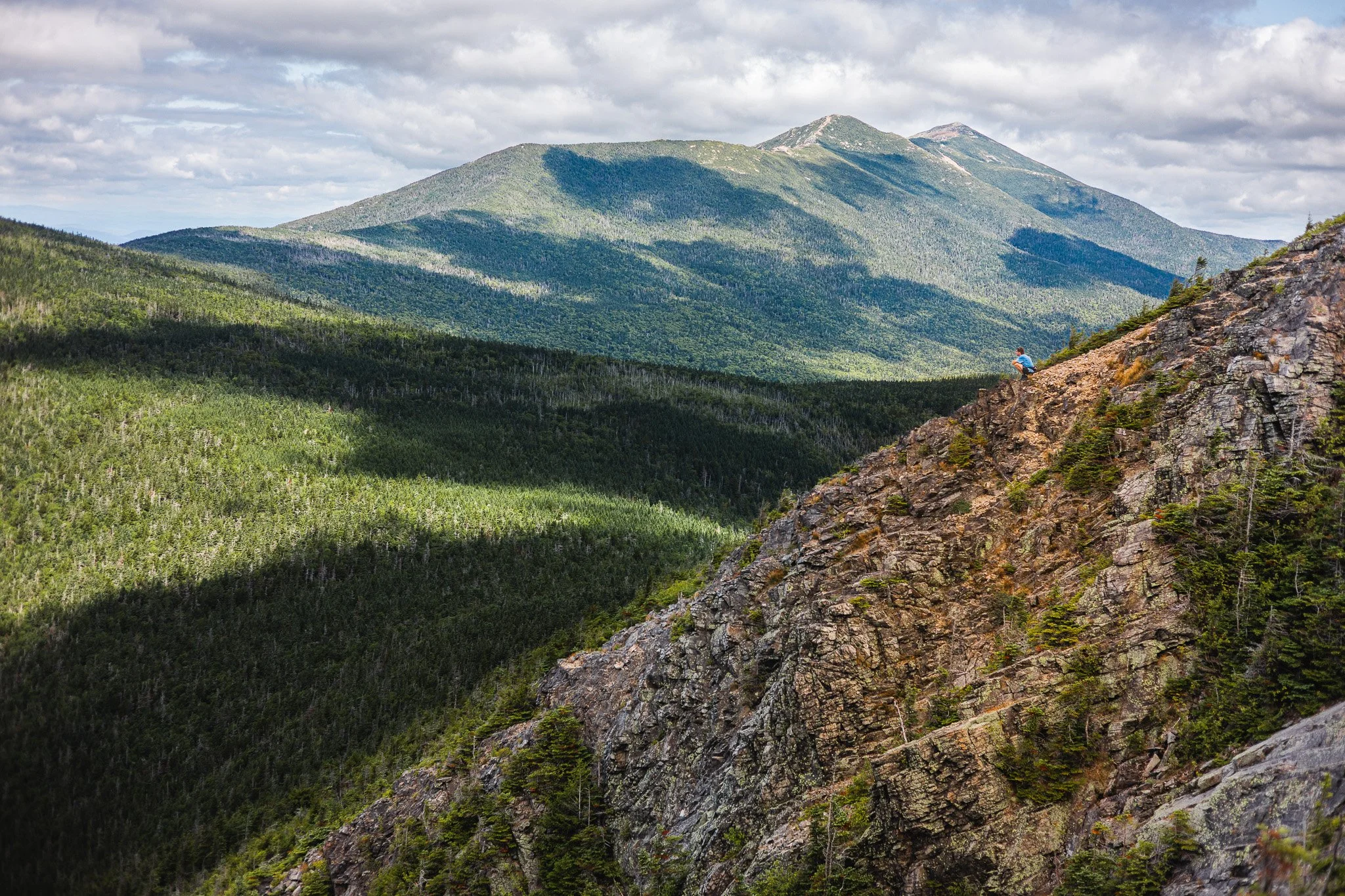 A hiker in a blue jacket sitting on a rocky slope overlooking a forested mountain landscape with a large mountain range in the background under a cloudy sky.