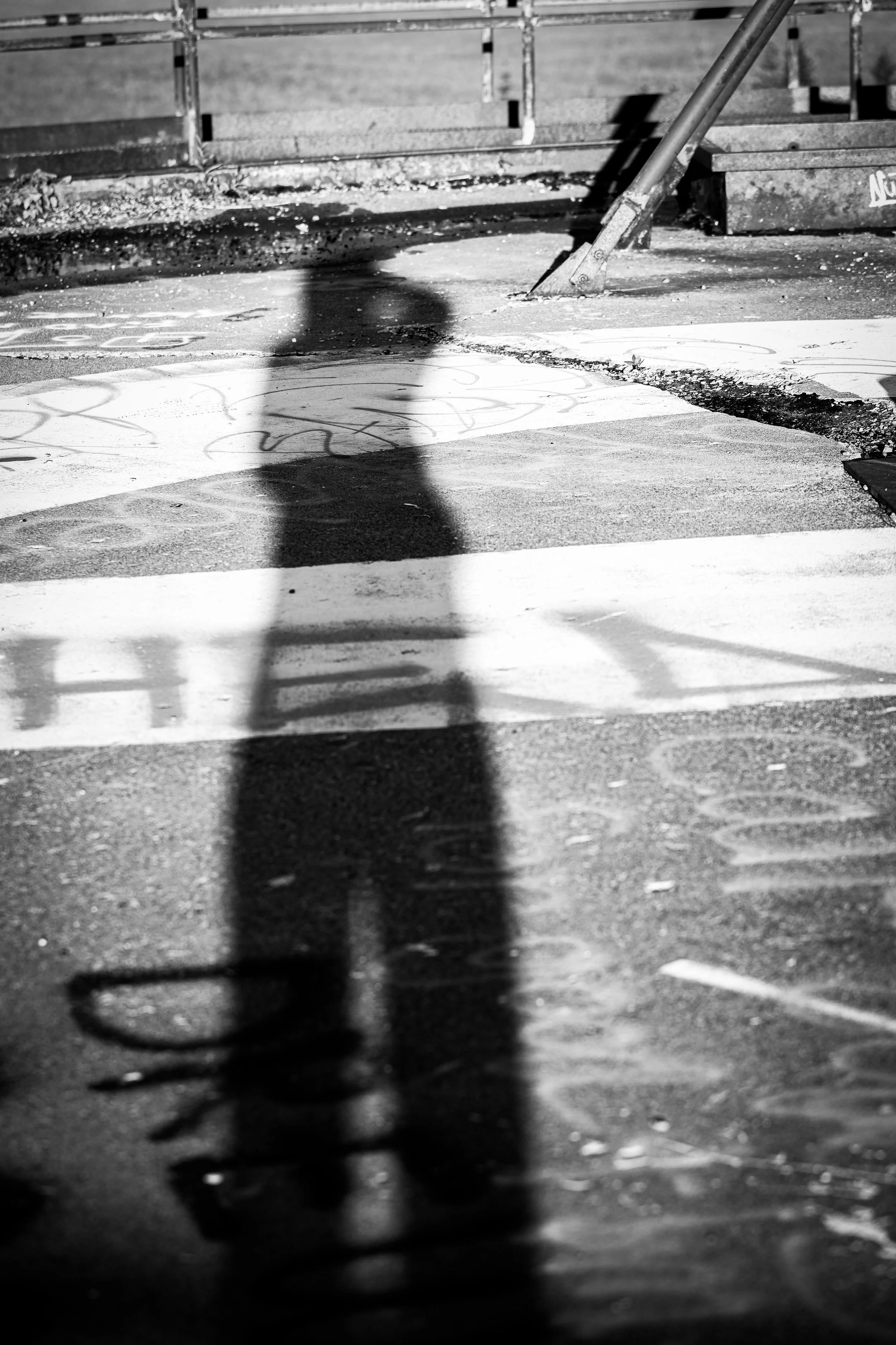 Black and white photo of a person’s shadow cast on a sidewalk, holding a bicycle handlebar, with graffiti on the ground and a barrier in the background.