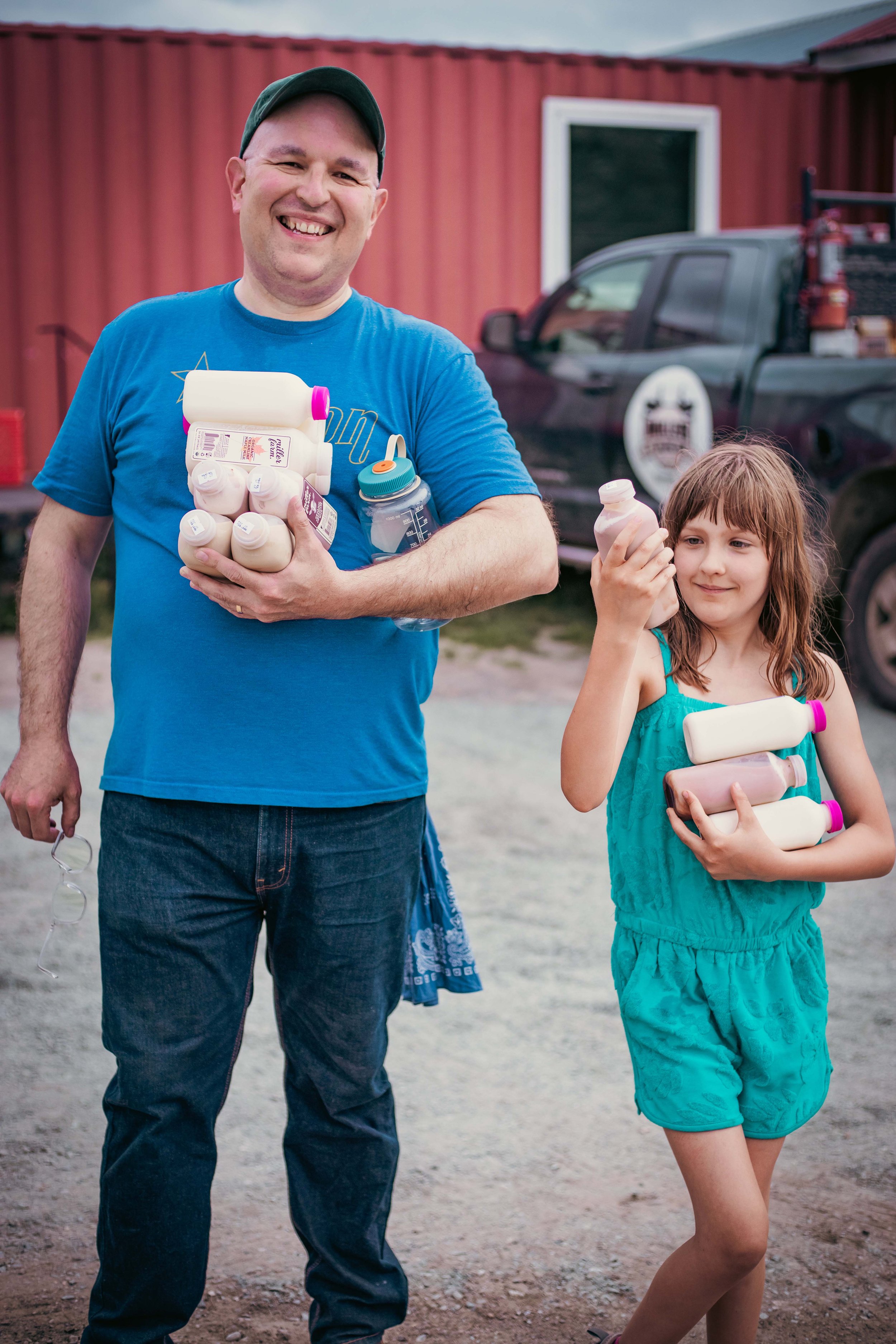A man and a young girl standing outdoors, each holding bottles of milk, smiling. The man is wearing a blue shirt and a green cap, and the girl is wearing a turquoise romper. In the background, there is a red building, a black pickup truck, and a cloudy sky.