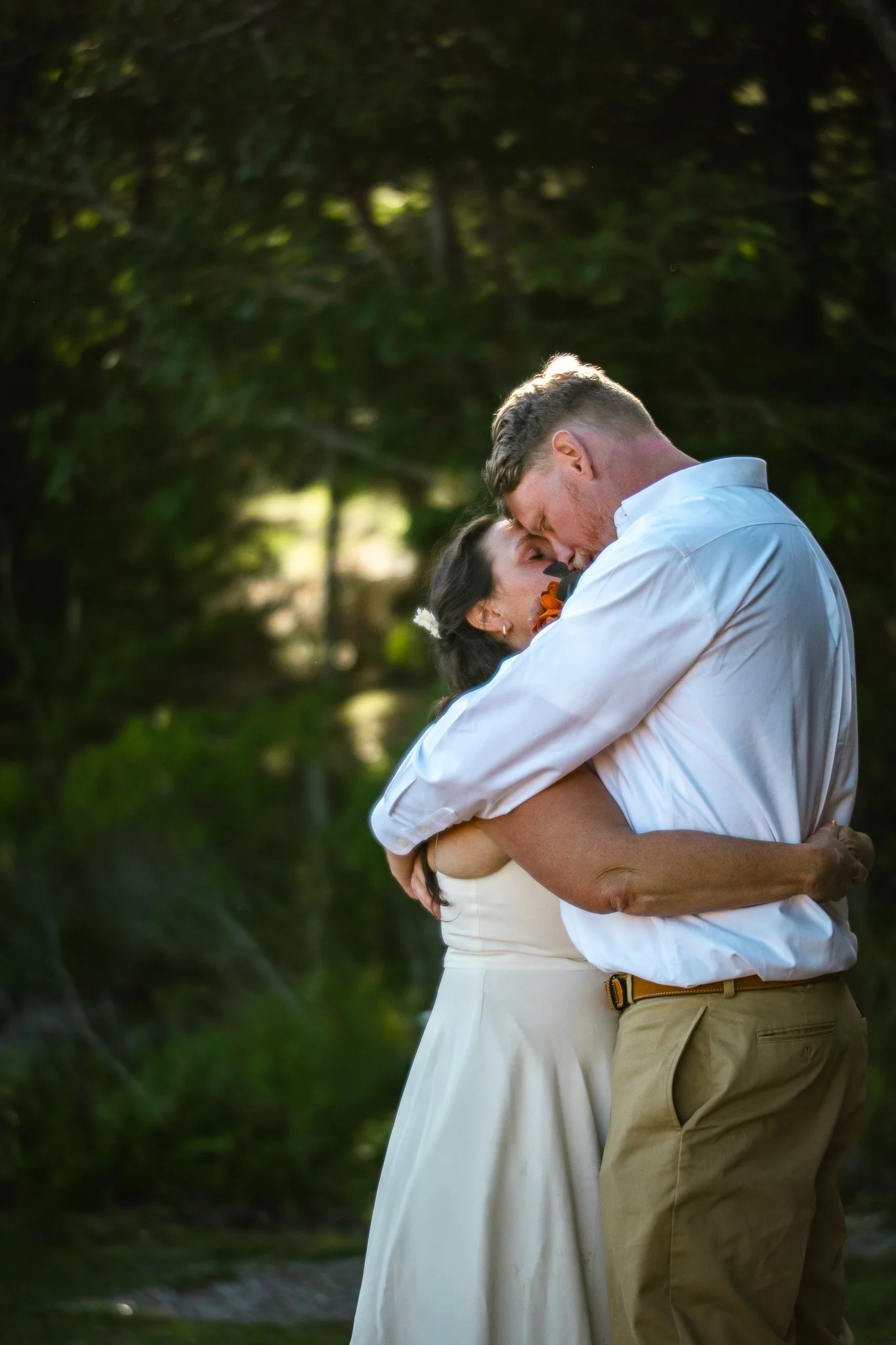 A couple is embracing outdoors, with the man dipping the woman as they hold each other close, surrounded by trees and natural greenery.