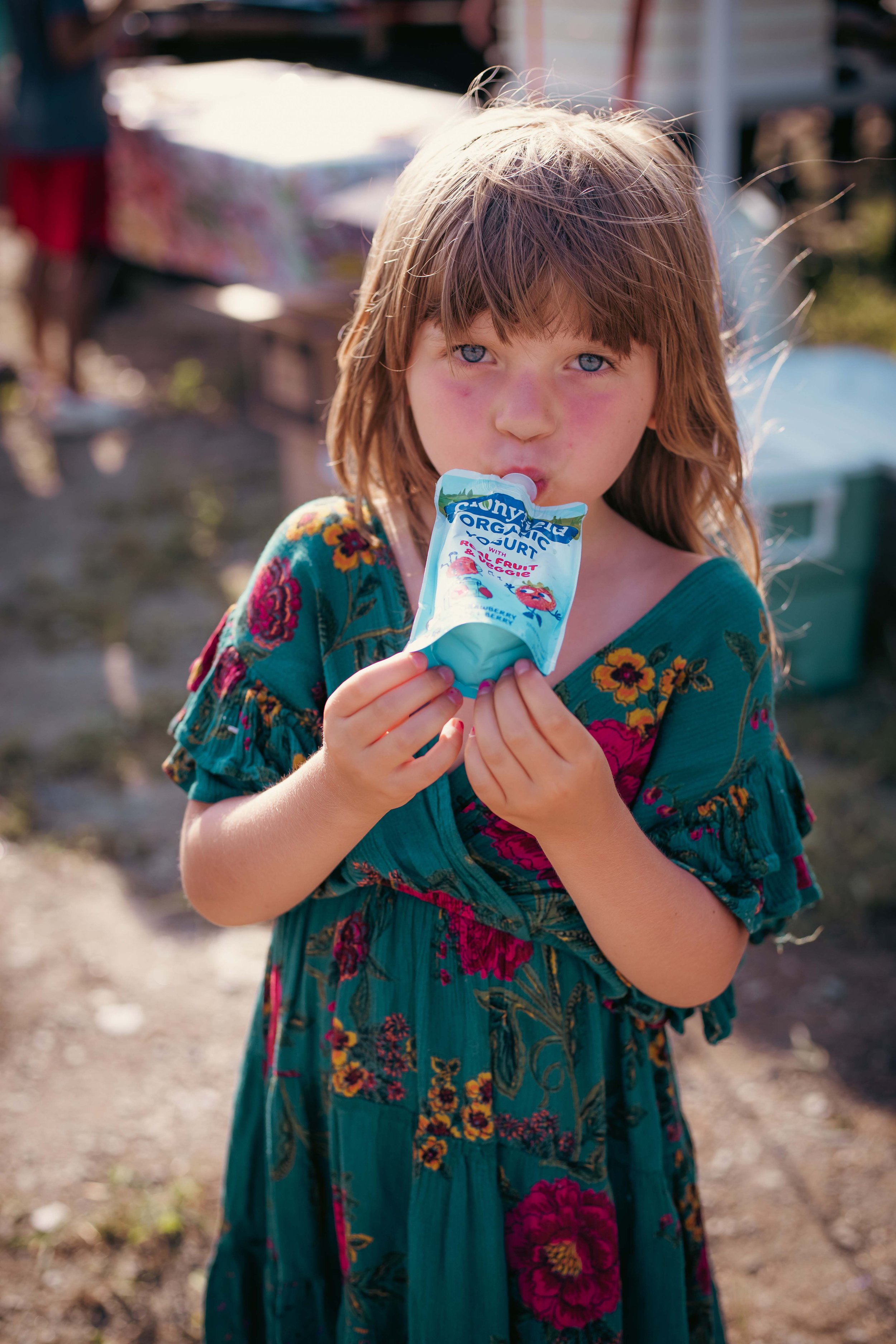 A young girl with blue eyes and brown hair wearing a colorful floral dress, holding a small container of yogurt with strawberries, outdoors with blurred background.