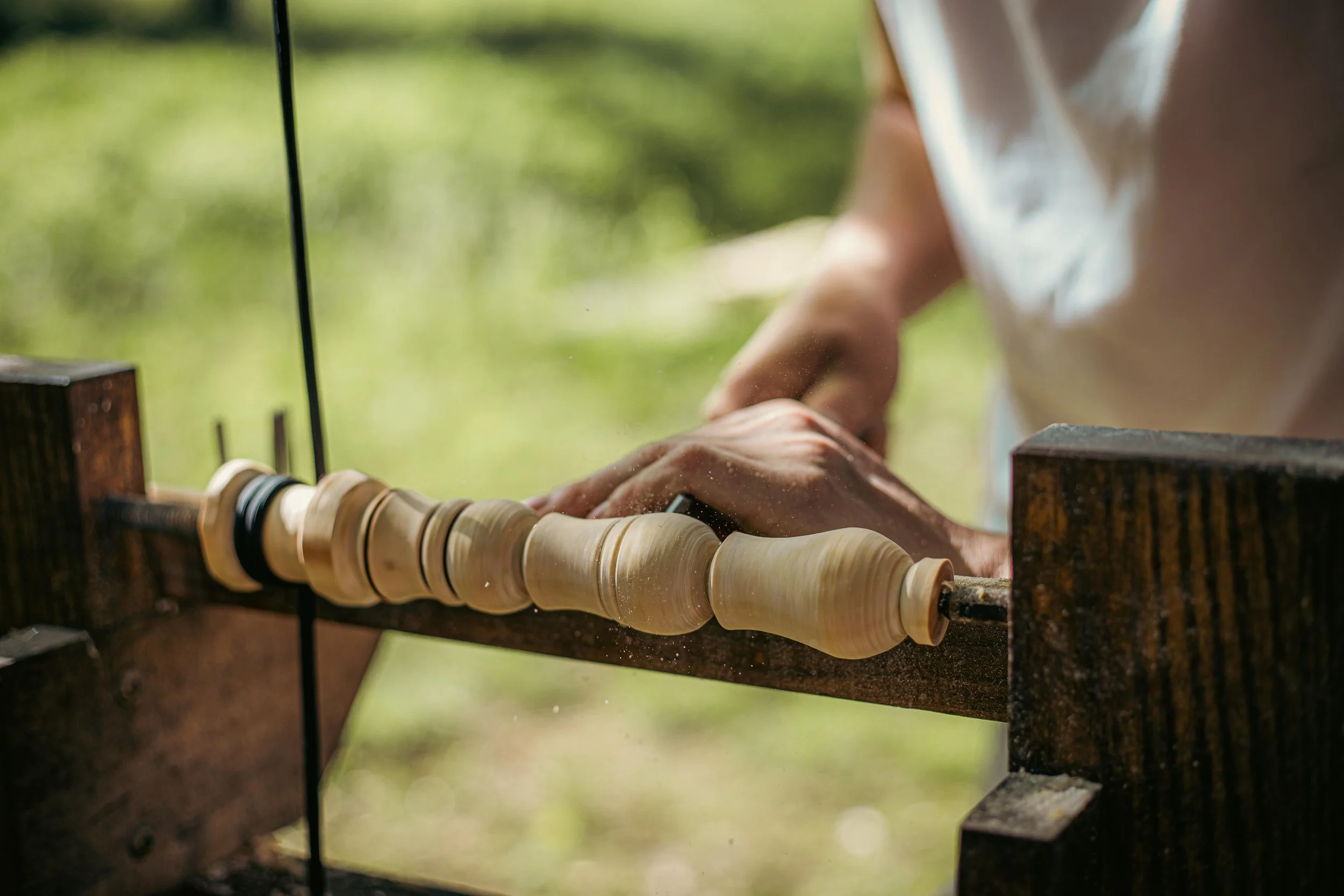 Person carving a wooden object on a lathe outdoors with greenery in the background.