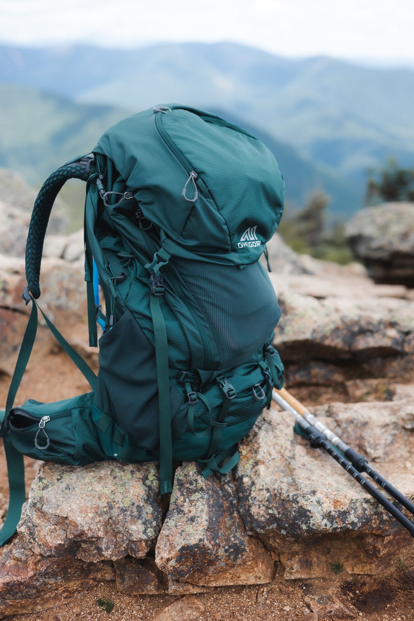 A teal hiking backpack on rocks in a mountainous landscape with misty mountains in the background, also a hiking pole leaning on the rocks.
