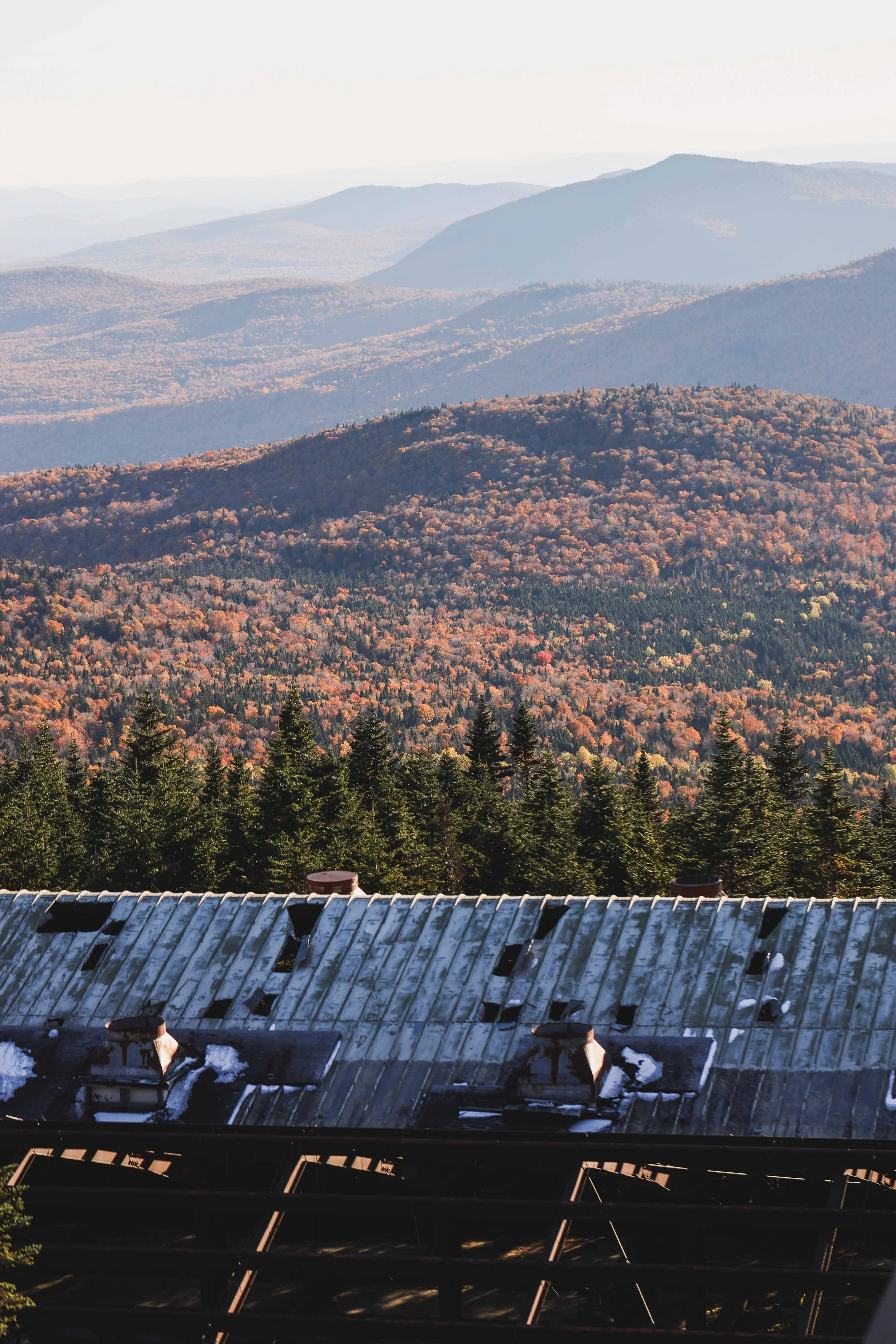 View from a building showing a forested mountain range with layers of rolling hills and mountains in the distance, under a bright sky. The foreground includes part of a metal roof with chimneys.