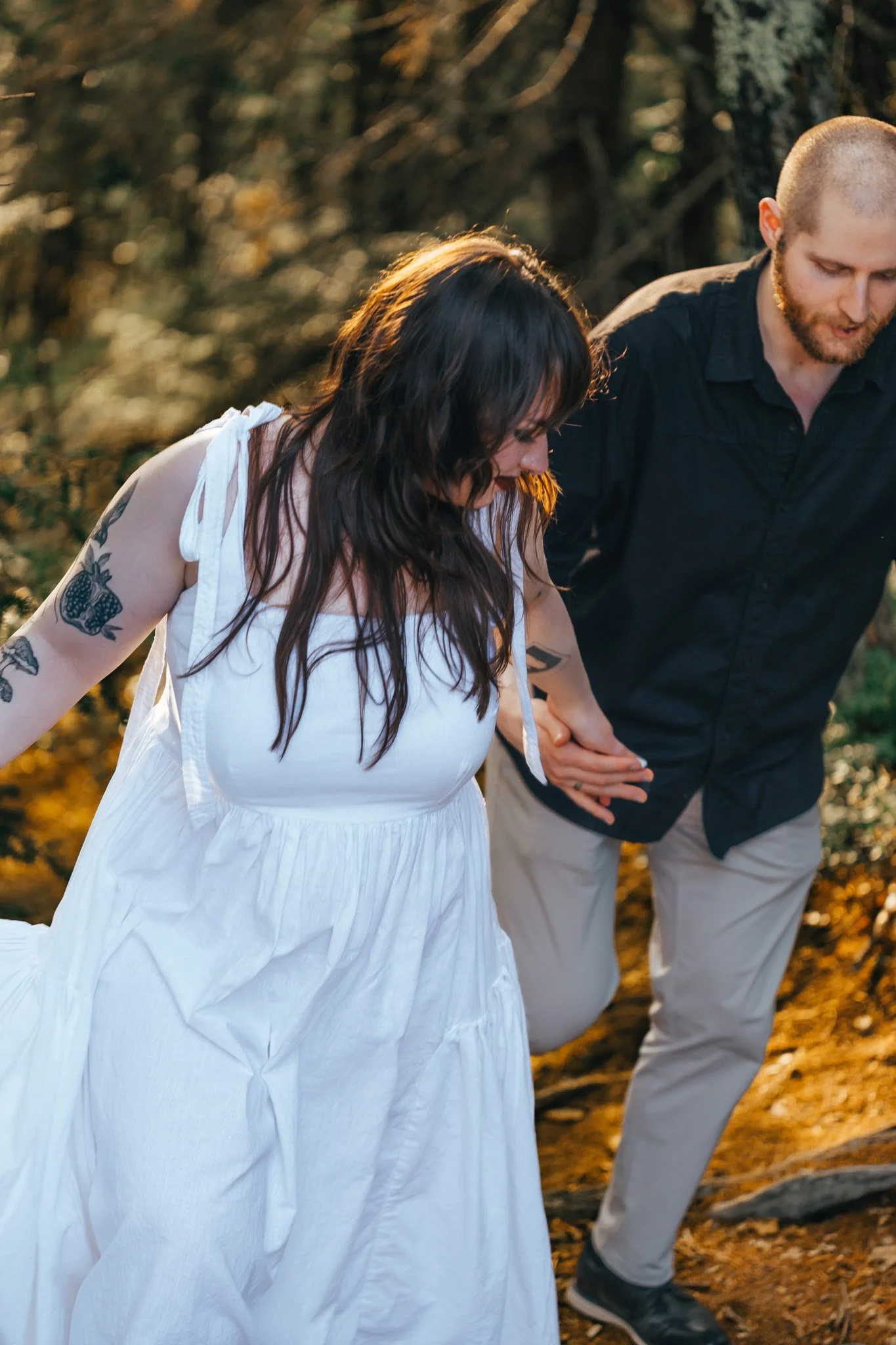 A woman and a man are holding hands and walking outdoors on a trail in the woods during the daytime. The woman is wearing a white dress, has long dark hair, and tattoos on her arm. The man has a beard, short hair, and is wearing a black shirt and light-colored pants.