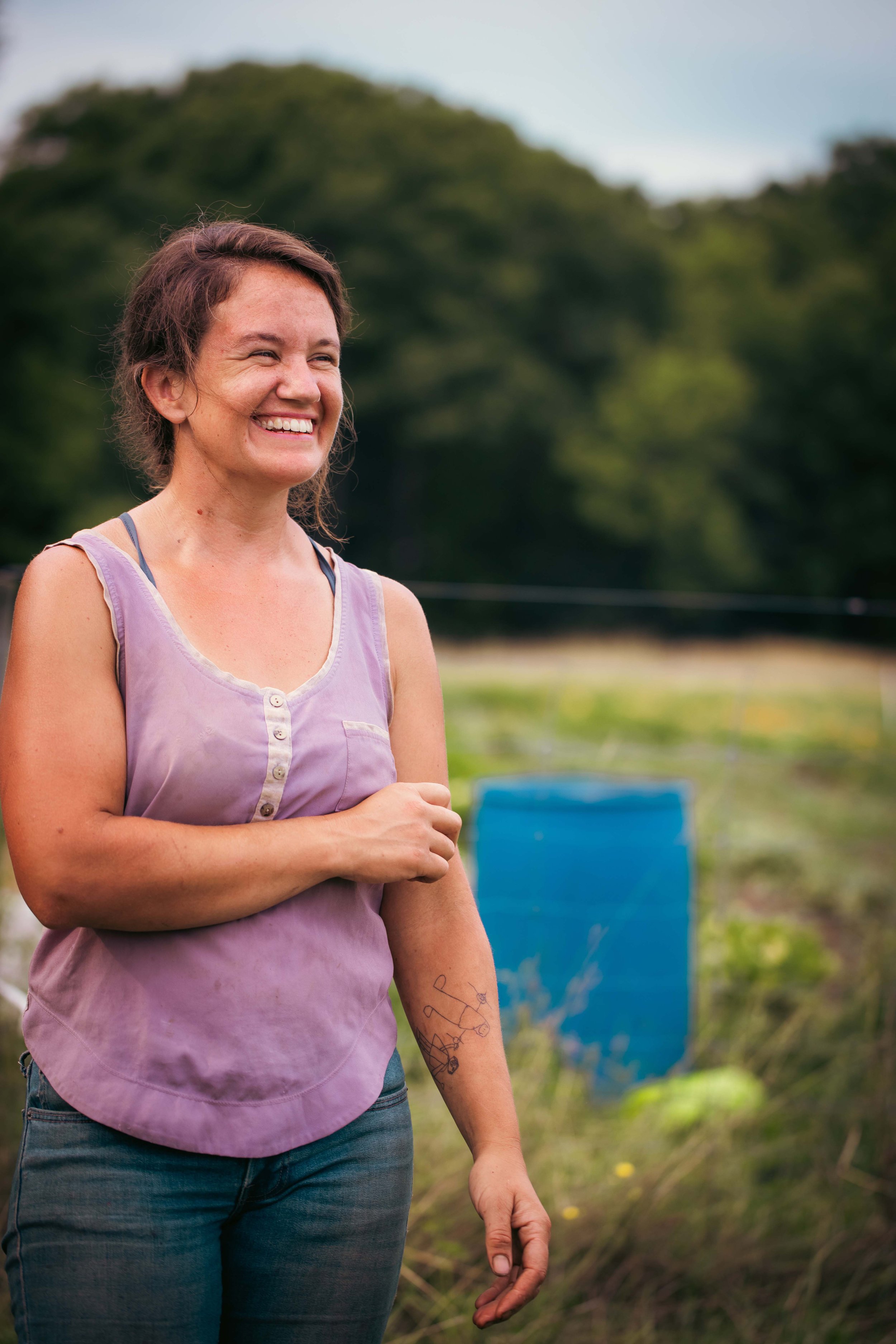 A woman smiling outdoors, wearing a purple sleeveless shirt, with a tattoo on her left forearm, standing in front of green trees and blue chairs.