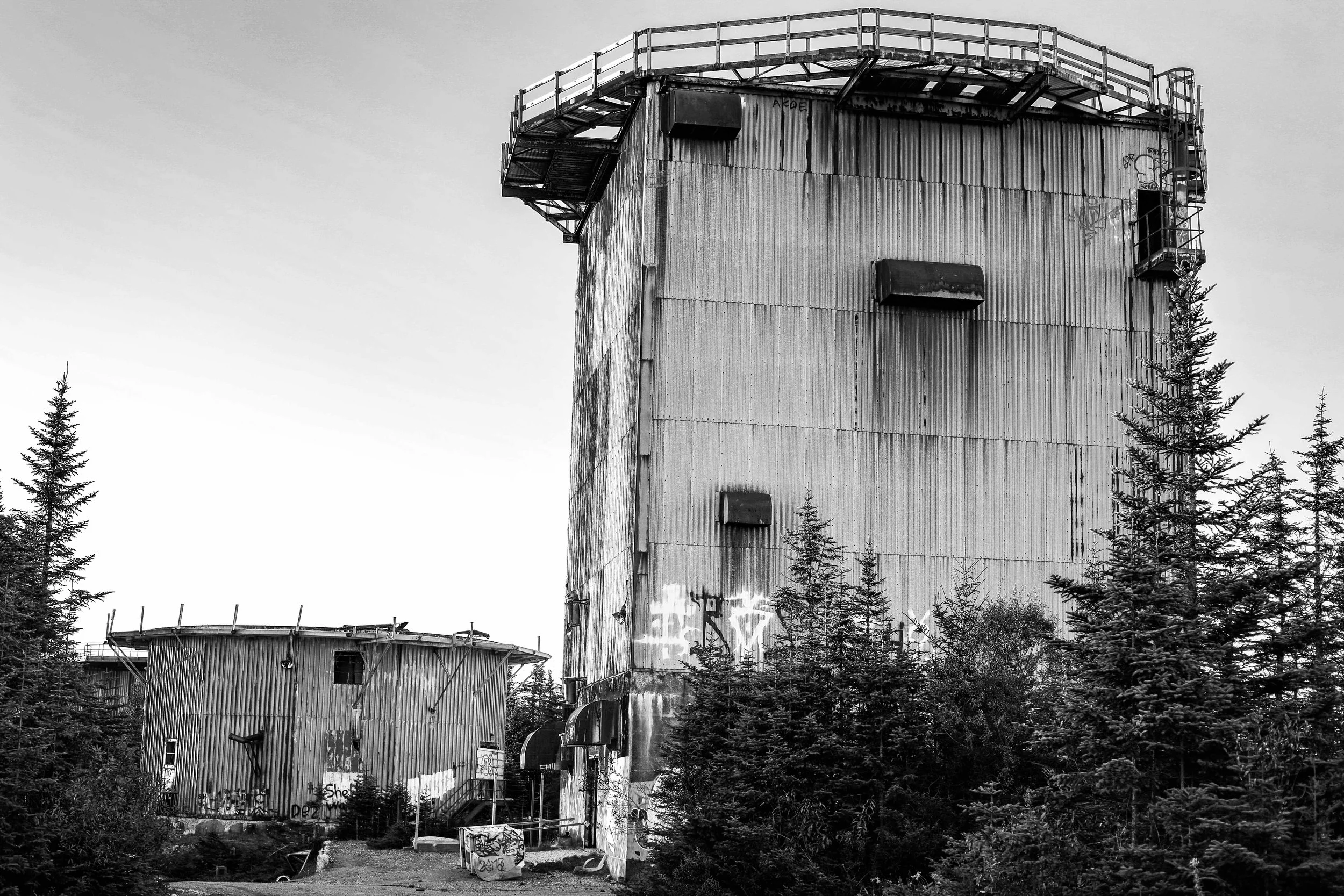 Black-and-white photo of an old, dilapidated industrial building surrounded by trees. The building has rusted metal siding and a rooftop balcony with a guardrail, with graffiti visible on the walls.