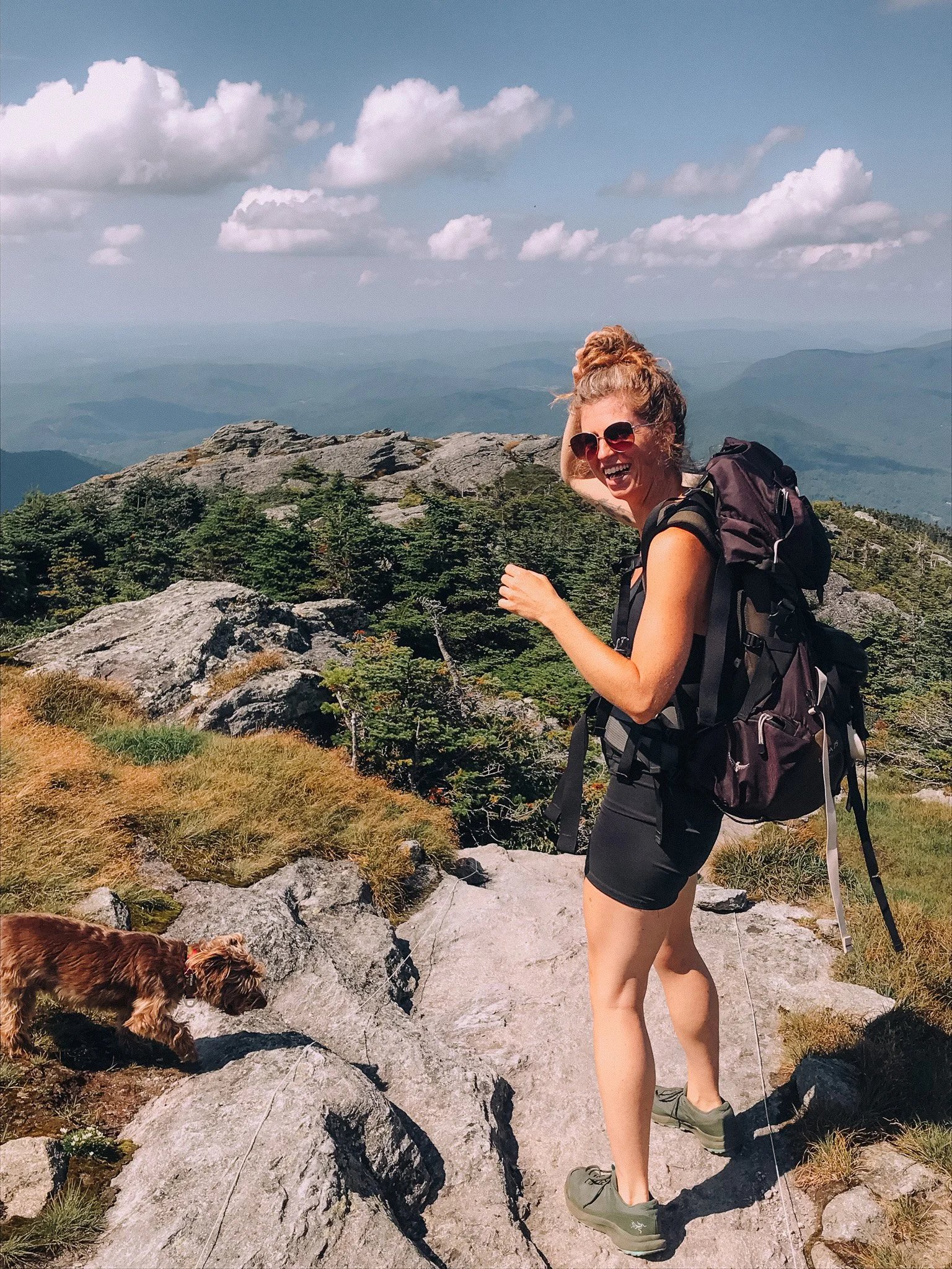 A woman with curly hair in a bun, wearing sunglasses, a black t-shirt, black shorts, and hiking shoes, is smiling and holding her hair back with one hand while hiking on rocky terrain with green bushes and trees, with mountain ranges and a partly cloudy sky in the background. A small brown dog is walking nearby on the trail.