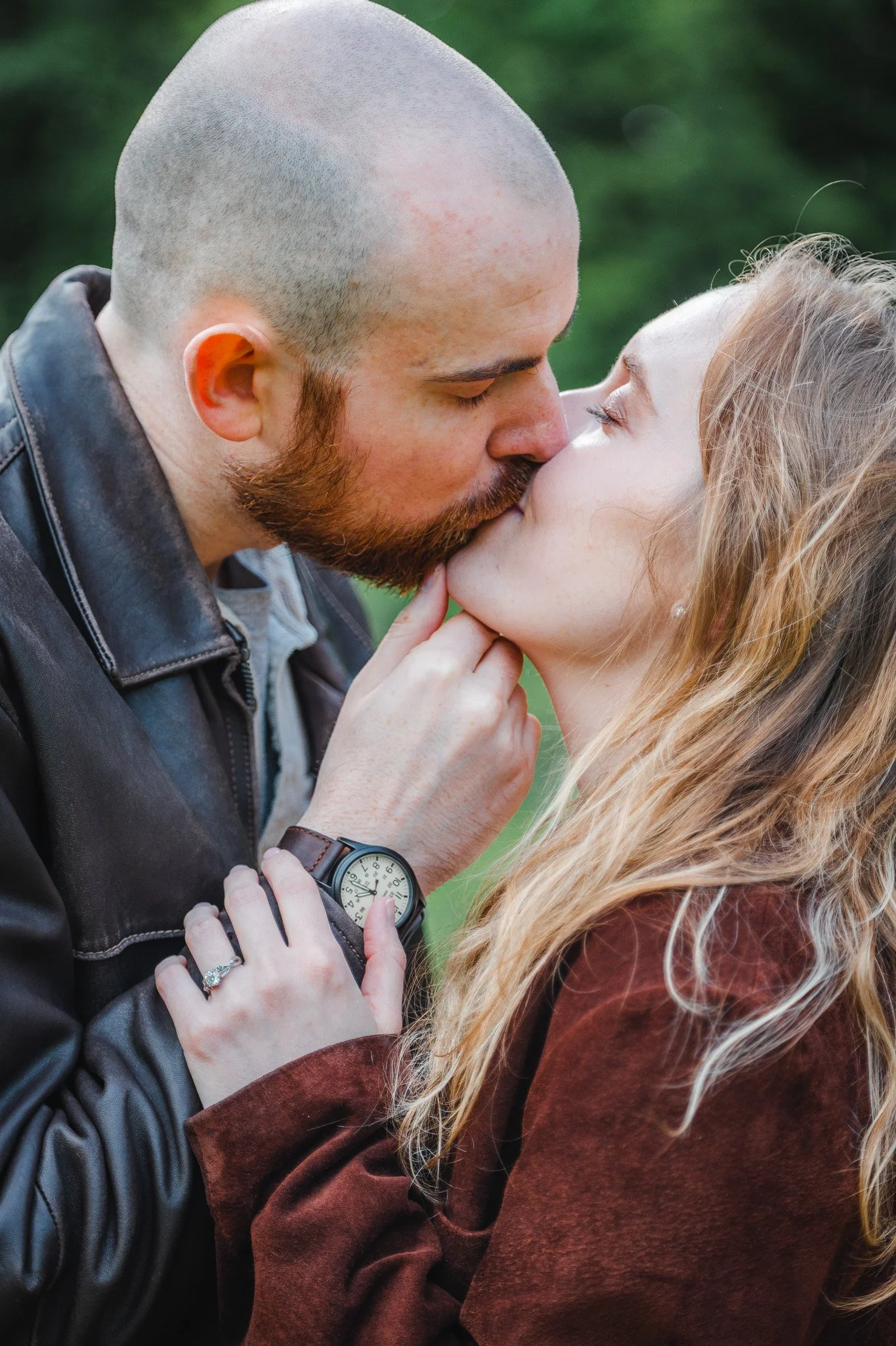 A couple kissing outdoors, with the woman wearing a ring and a watch, and the man wearing a leather jacket.