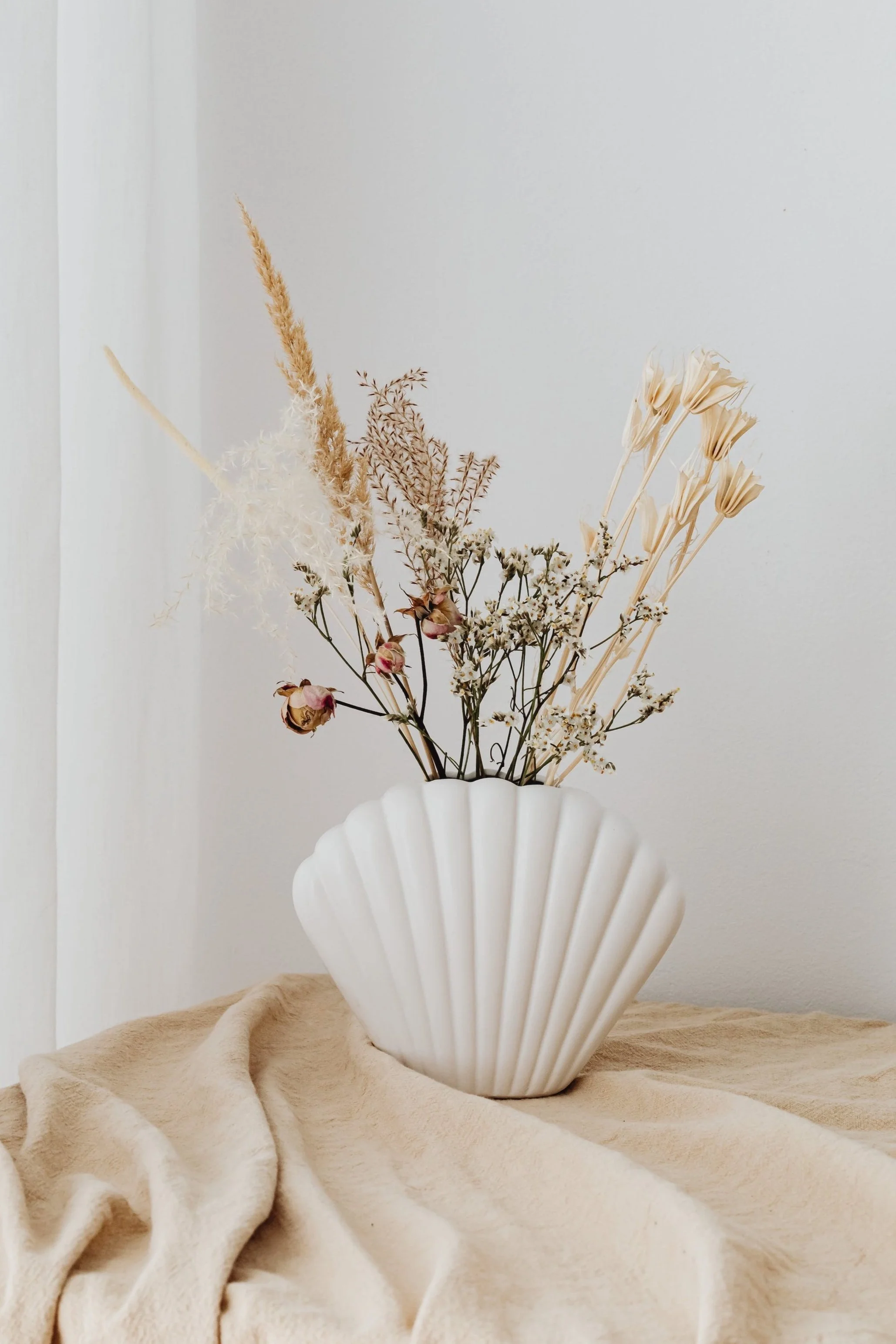 A white, scalloped-shell-shaped ceramic vase holding dried flowers, including pampas grass, and placed on a beige fabric surface against a plain, light-colored wall.