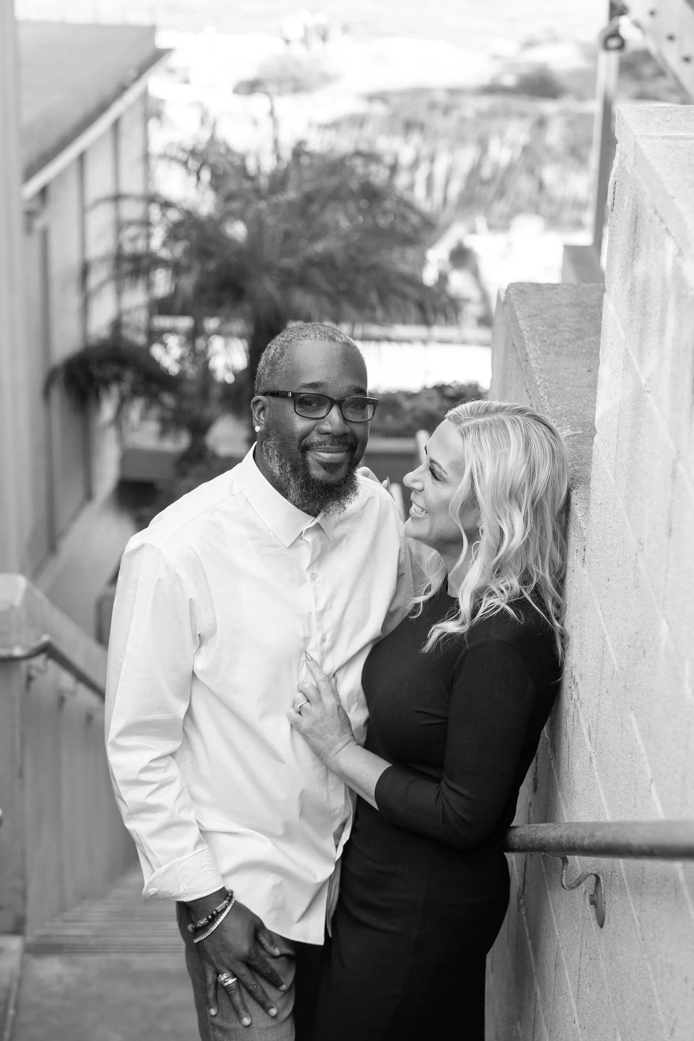 A Black man with glasses and a beard stands outdoors next to a blonde woman with wavy hair, who is smiling at him. They are close together, with the woman holding his hand and leaning against a wall, smiling at him. The background includes trees and a railing.