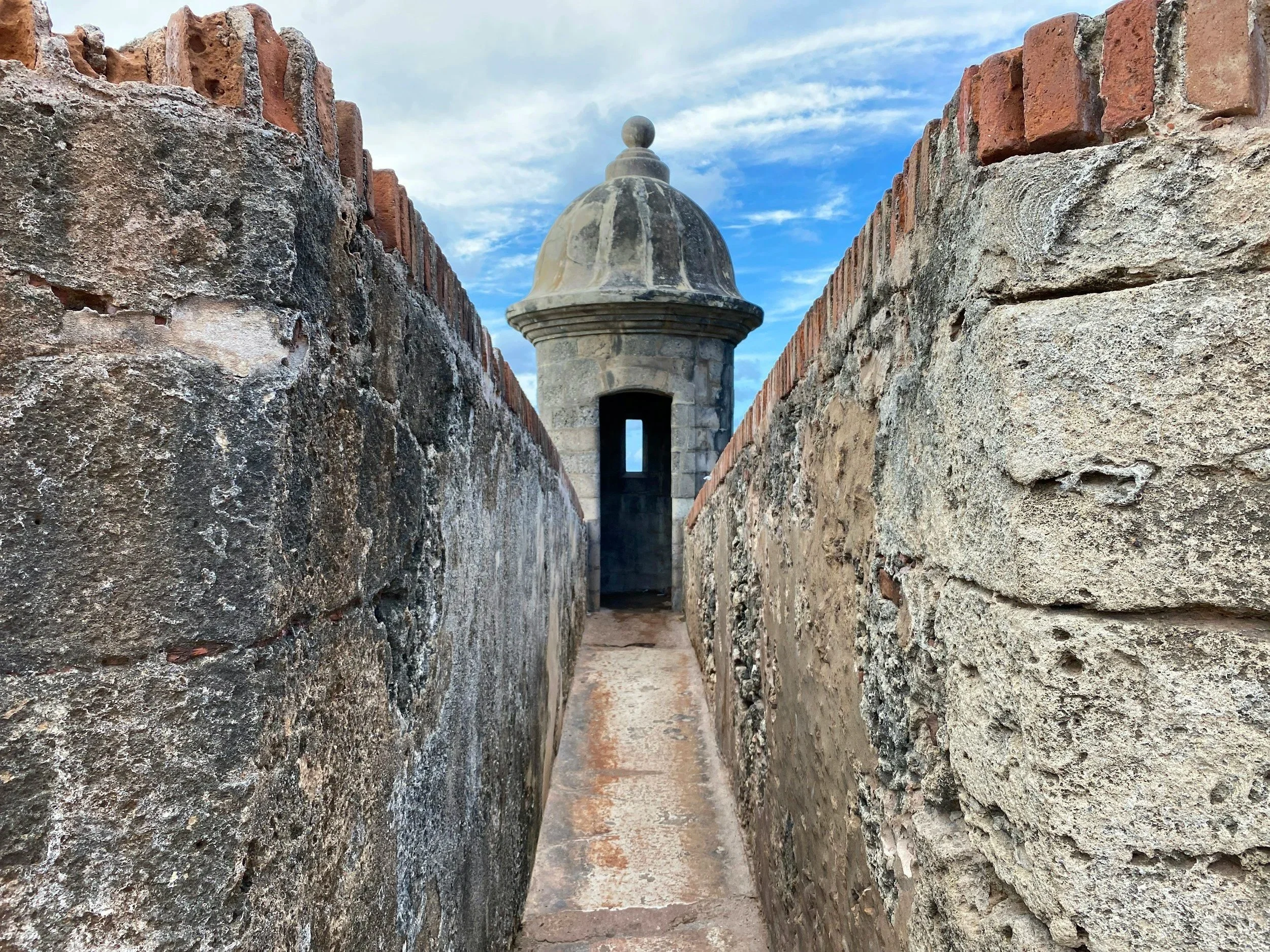 A narrow passageway between two textured stone walls leading to a small arch with a domed top and a small window, against a partly cloudy sky.