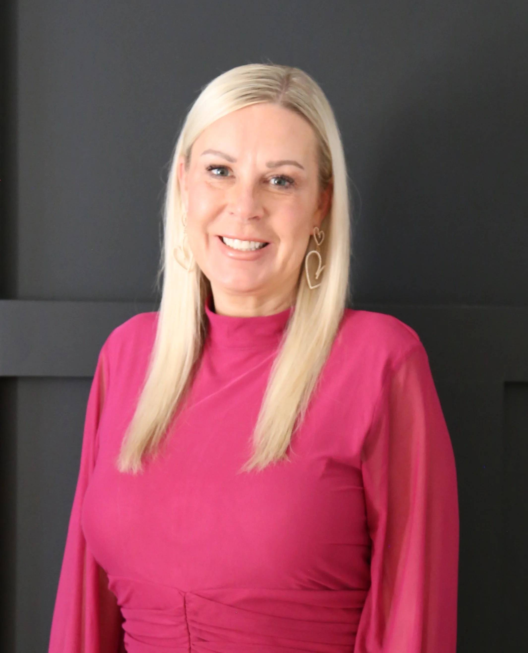 Close-up image of a woman with blonde hair, wearing a pink top and decorative earrings, smiling in front of a dark background.