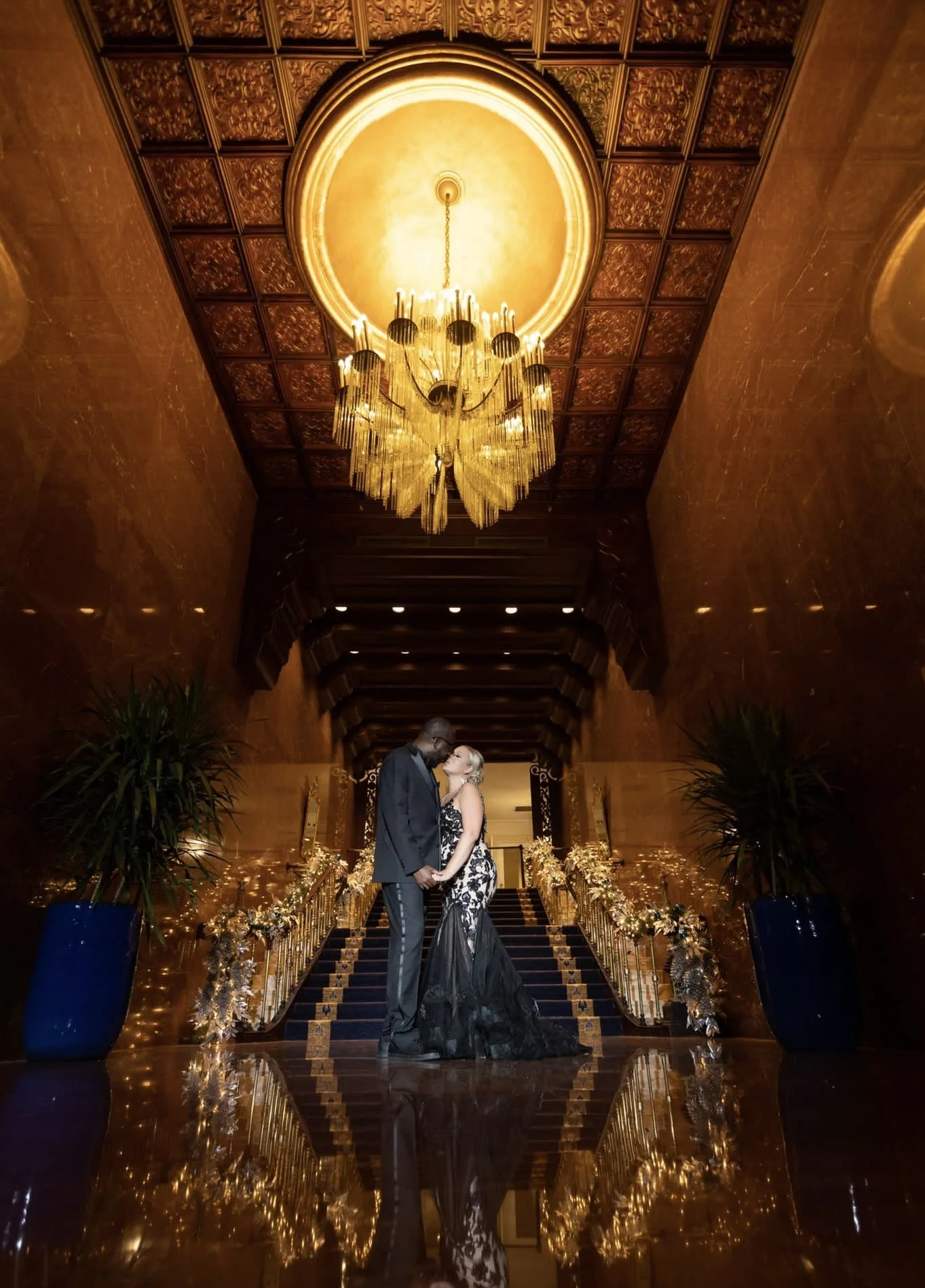 A couple dressed in elegant black and white formal wear, holding hands and gazing into each other's eyes, standing on a staircase decorated with white flowers, Christmas lights, and gold accents, in a grand indoor setting with a chandelier and ornate ceiling.