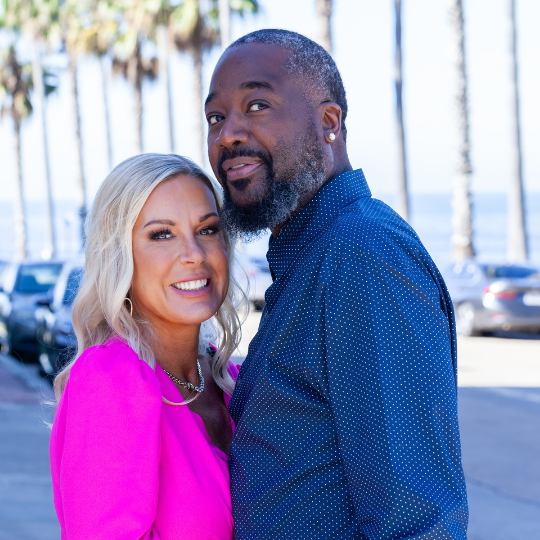 A smiling blonde woman in a pink top standing next to a dark-skinned man in a blue shirt, with a background of palm trees and parked cars.